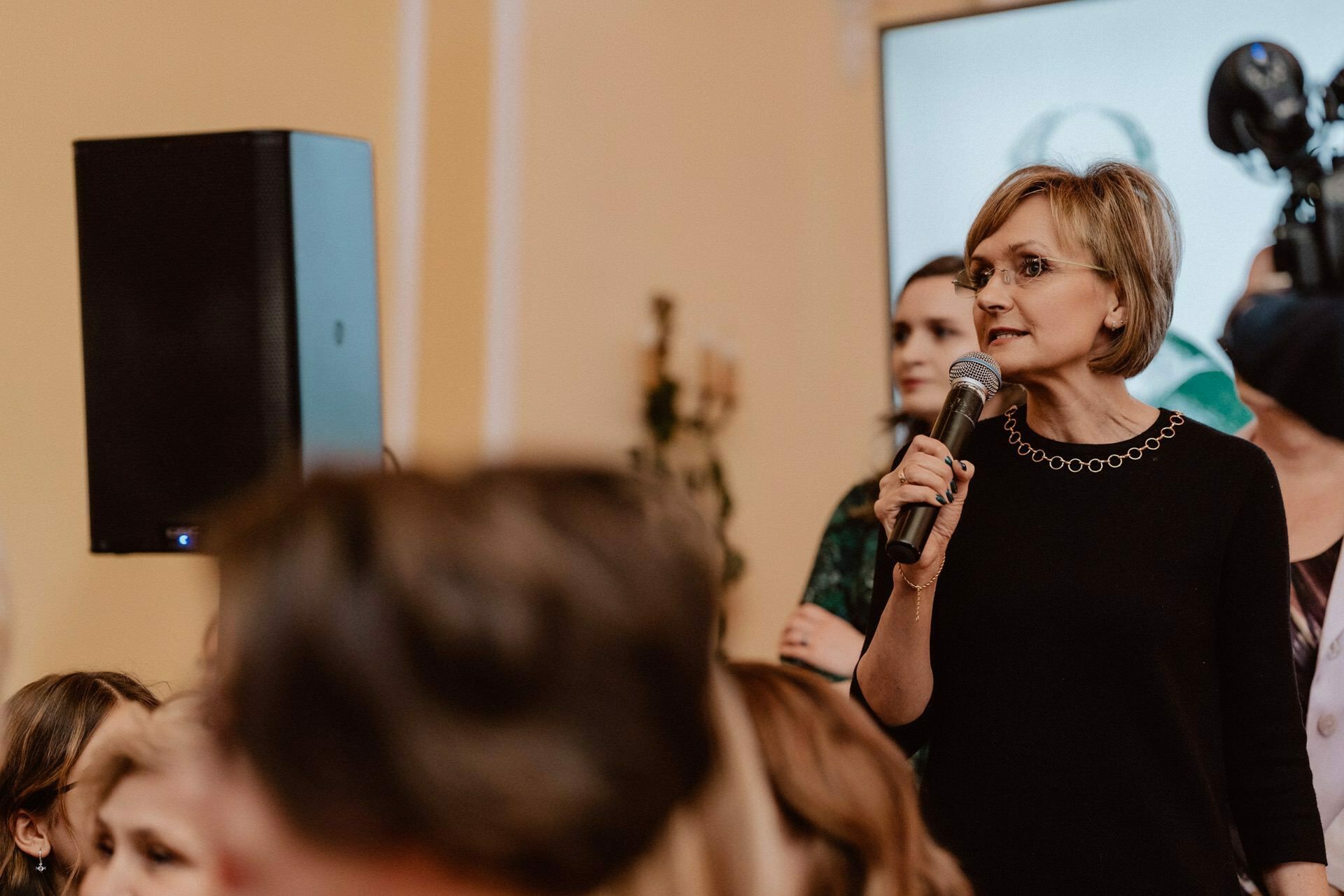 A woman with short hair and glasses speaks into a microphone in front of a crowd. She is standing in a room with beige walls, delivering what appears to be a report from a prom, with a large speaker behind her. Other people can be seen in the background, including someone with a camera.  