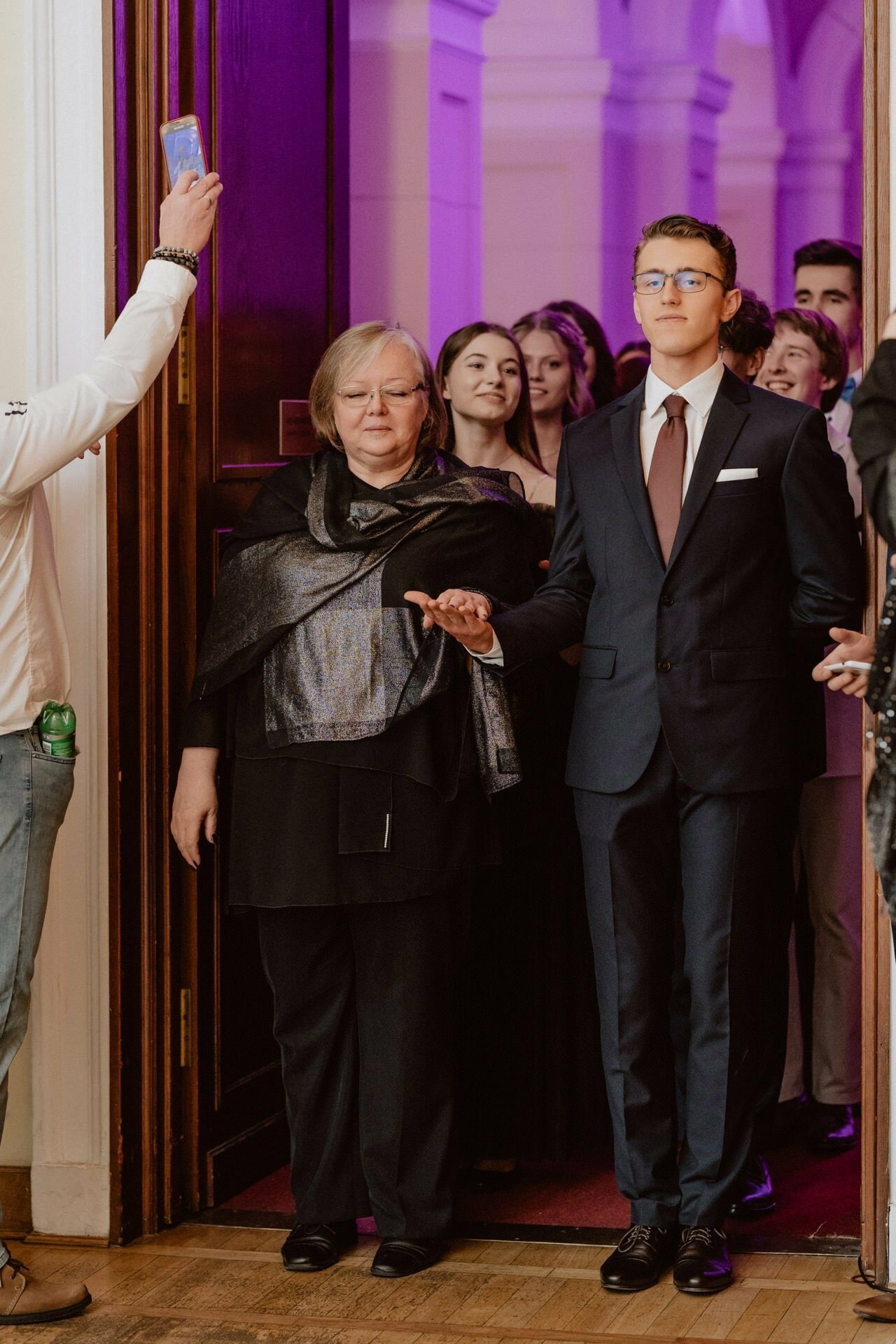 A woman and a young man, both dressed formally, enter a room filled with people. The woman is dressed in a black outfit with a shimmering scarf, while the man is in a dark suit and tie. More formally dressed people can be seen in the background, reminiscent of a reportage from a prom.  