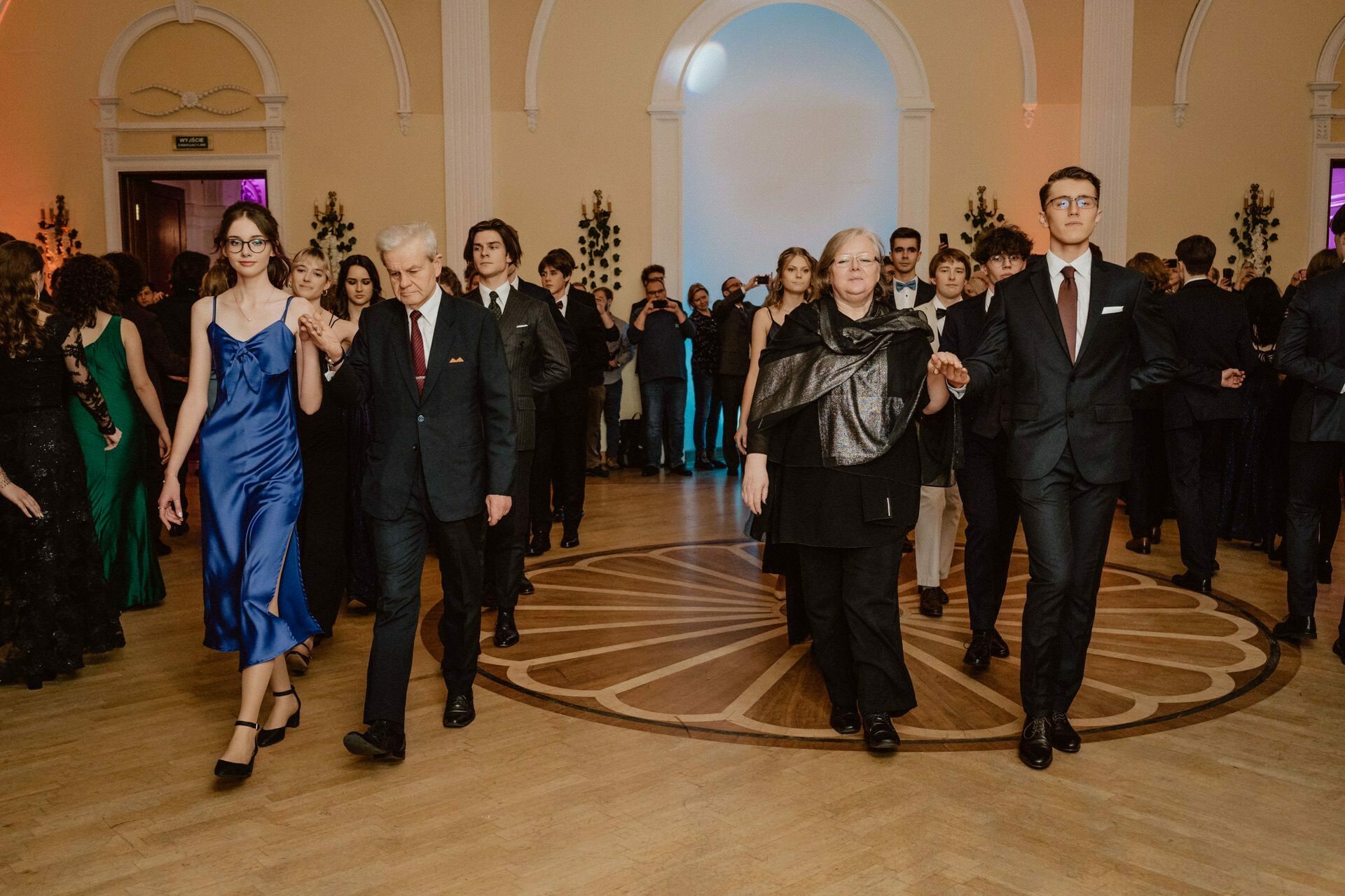 A group of people in formal attire are dancing in a large room with arched doors and a wooden floor, reminiscent of a prom reportage. Two couples are in the foreground, holding hands and walking. Other guests are seen in the background, mingling and dancing. The atmosphere is elegant and festive.   