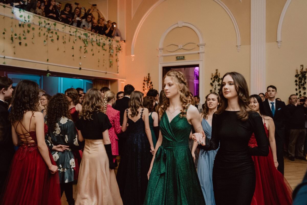 W wielkim, eleganckim pokoju z wysokimi sufitami i ozdobnymi dekoracjami spotyka się grupa osób ubranych w formalne stroje. W centrum reportażu ze studniówki wyróżniają się dwie młode kobiety, jedna w zielonej, a druga w czarnej sukience, trzymając się za ręce i wygrzewając się pod spojrzeniem balkonu.