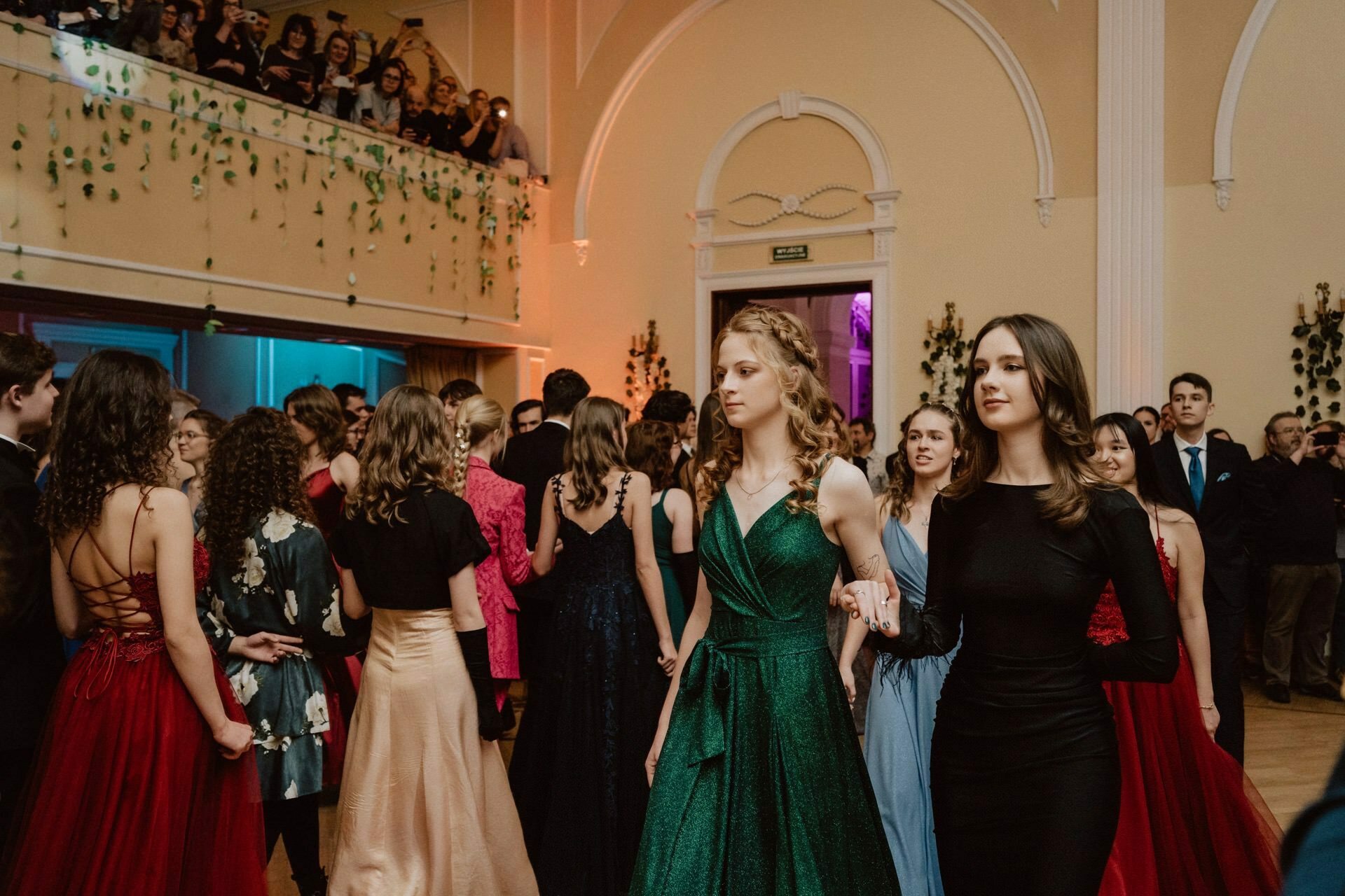 In a large, elegant room with high ceilings and ornate decorations, a group of people dressed in formal attire meet. At the center of the prom reportage stand out two young women, one in a green and the other in a black dress, holding hands and basking under the gaze of the balcony. 