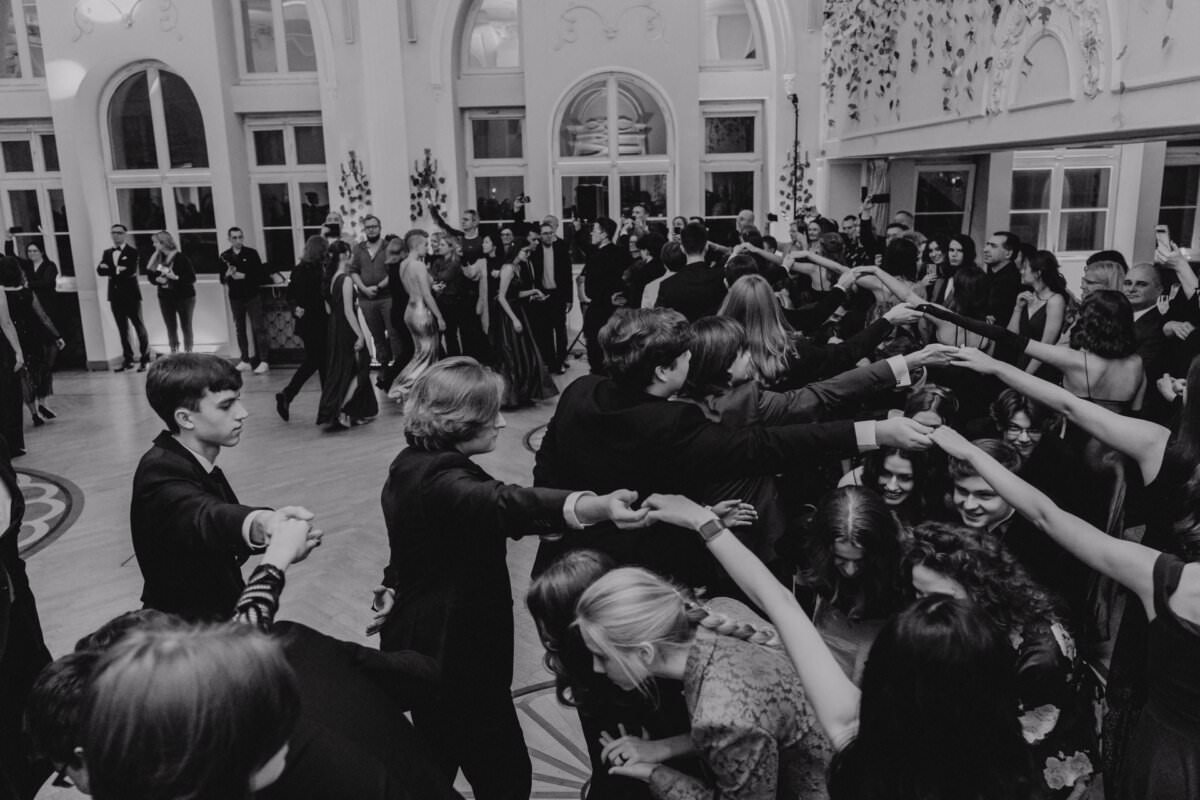 Grupa formalnie ubranych osób bierze udział w tańcu towarzyskim w bogato zdobionej sali, przypominającej reportaż ze studniówki. Niektórzy trzymają się za ręce w kręgu, inni patrzą. Przestrzeń ozdobiona jest łukowymi oknami i roślinami, emanując elegancką i świąteczną atmosferą. Obraz jest czarno-biały.