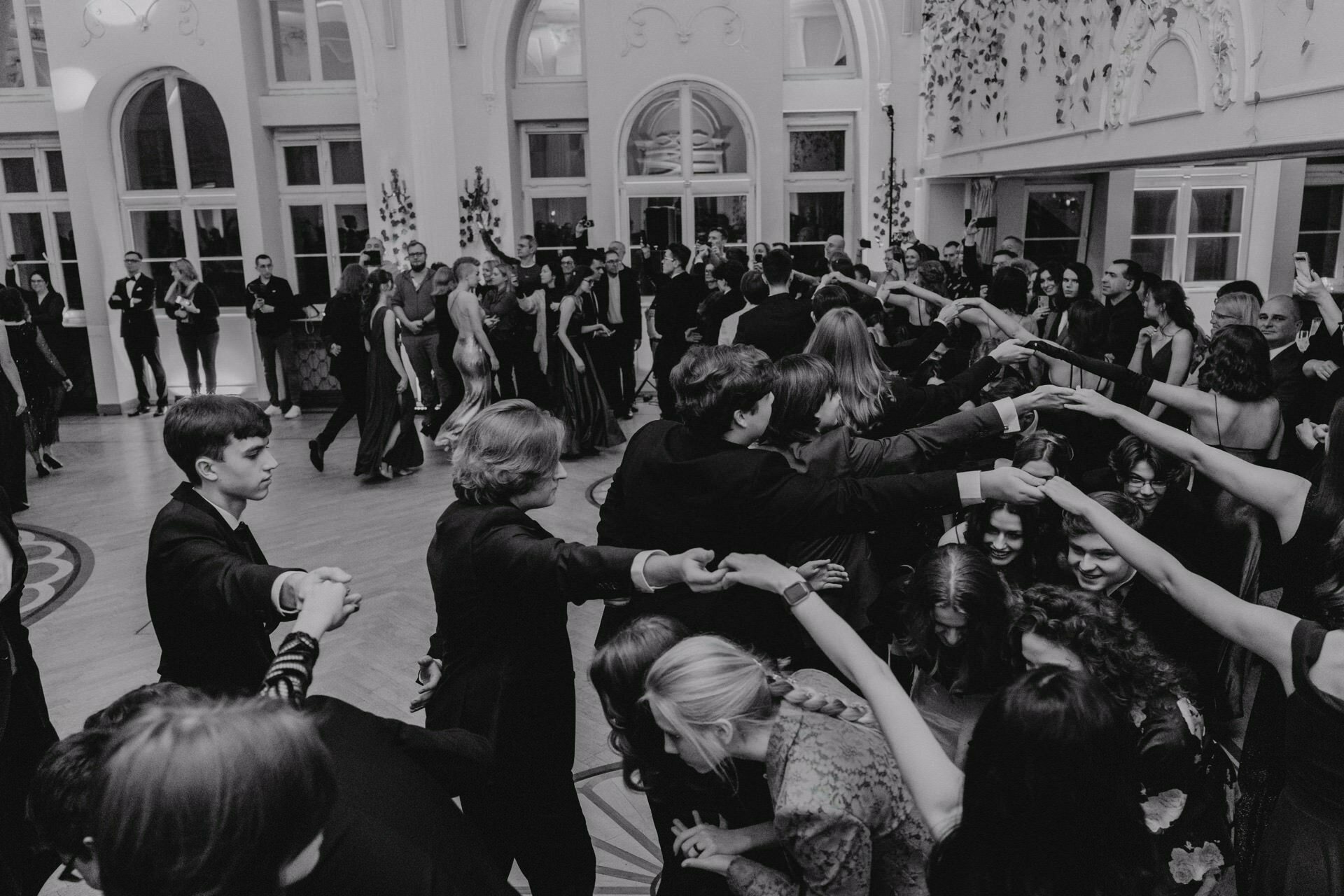 A group of formally dressed people take part in a ballroom dance in a richly decorated room, reminiscent of a prom report. Some hold hands in a circle, while others look on. The space is decorated with arched windows and plants, exuding an elegant and festive atmosphere. The image is in black and white.   