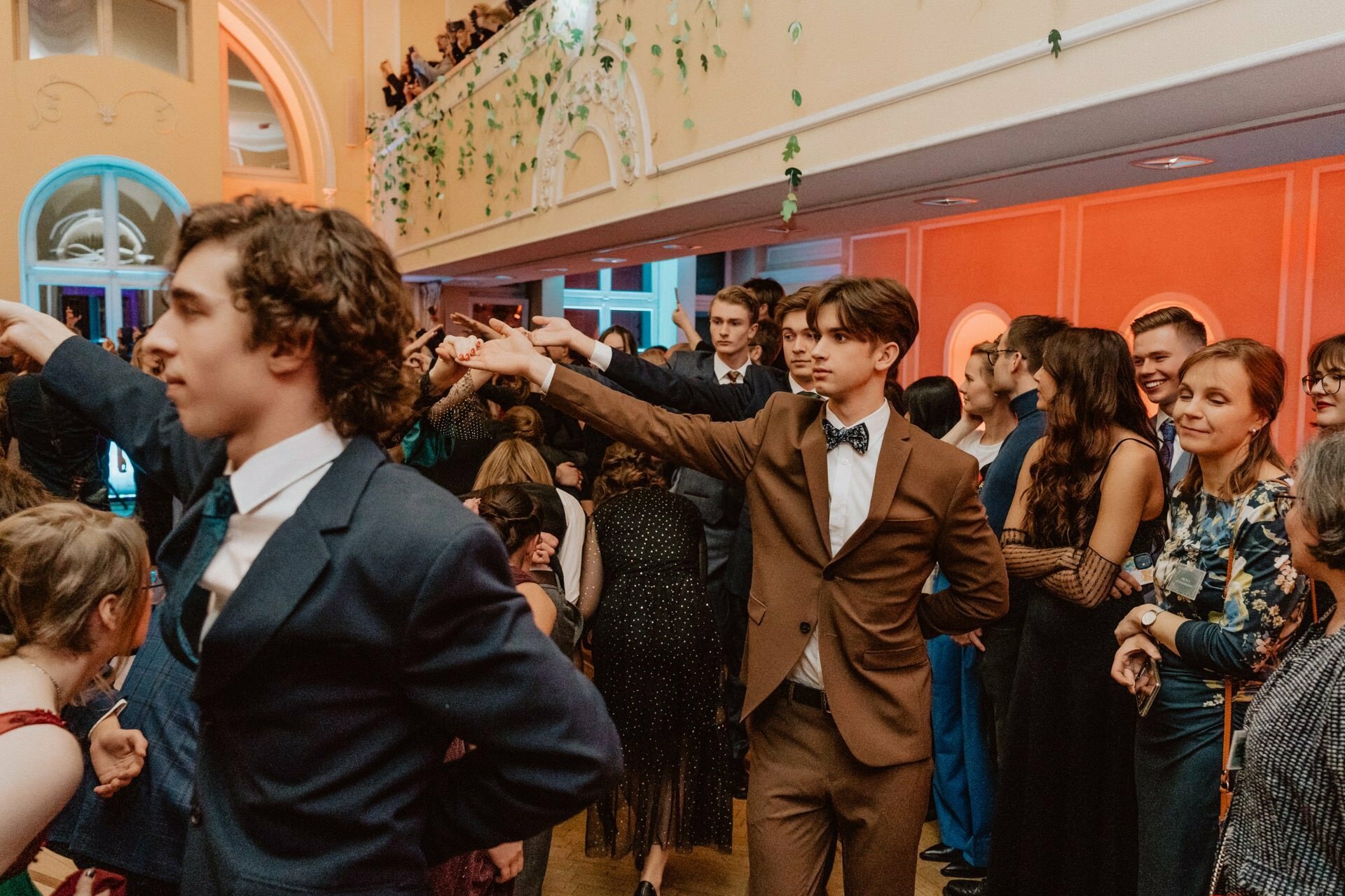 A lively ballroom scene reminiscent of a prom reportage with young couples dancing in formal attire. The central image focuses on two young men in suits, one in brown and the other in navy blue, holding hands with their female partners. Spectators line up in the background, enjoying the festive atmosphere.  