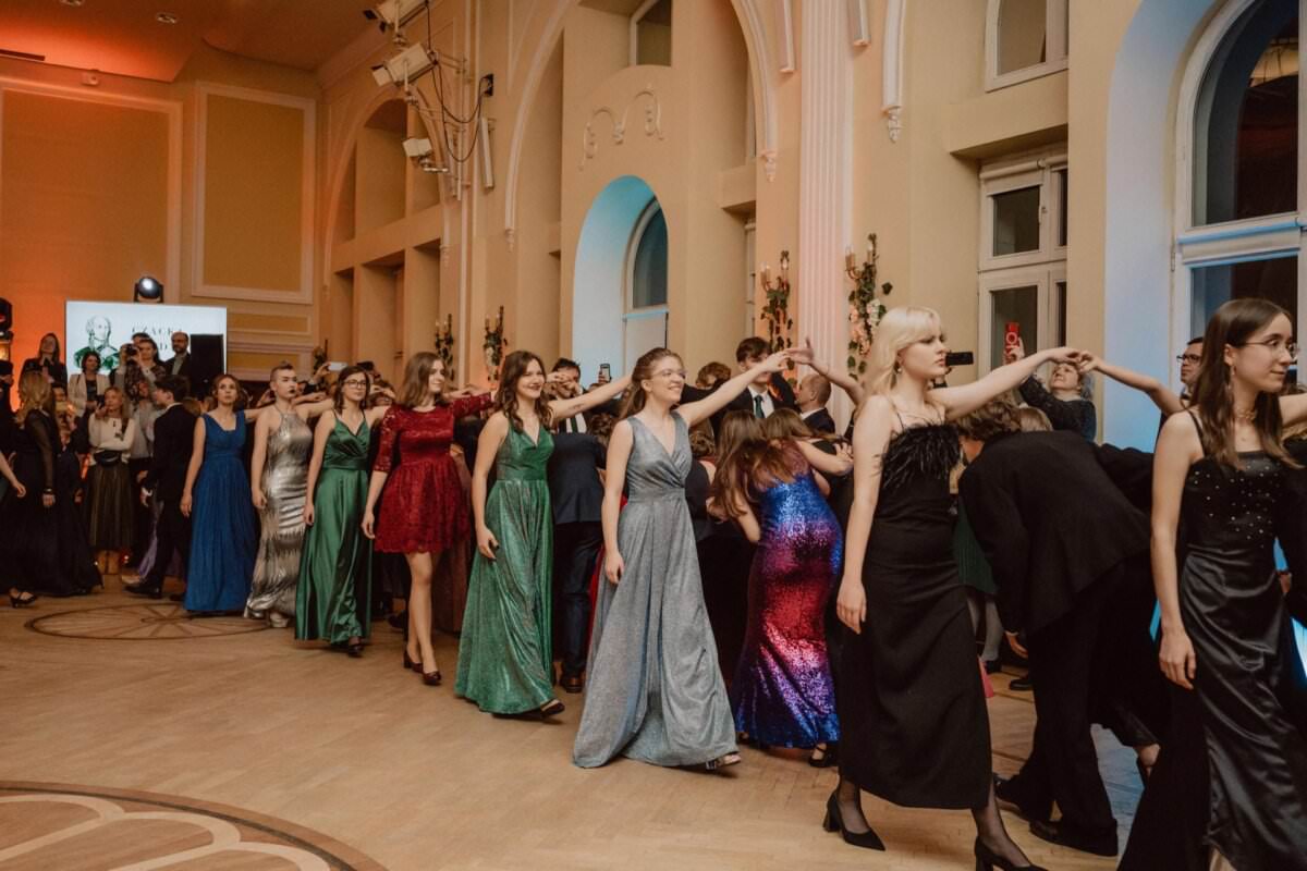 Grupa osób ubranych w formalne suknie wieczorowe i garnitury bierze udział w tańcu na imprezie w sali balowej. Ustawiają się w rzędzie conga, trzymają się za ręce i uśmiechają się, tańcząc w elegancko udekorowanej sali, przypominającej tętniący życiem reportaż ze studniówki.