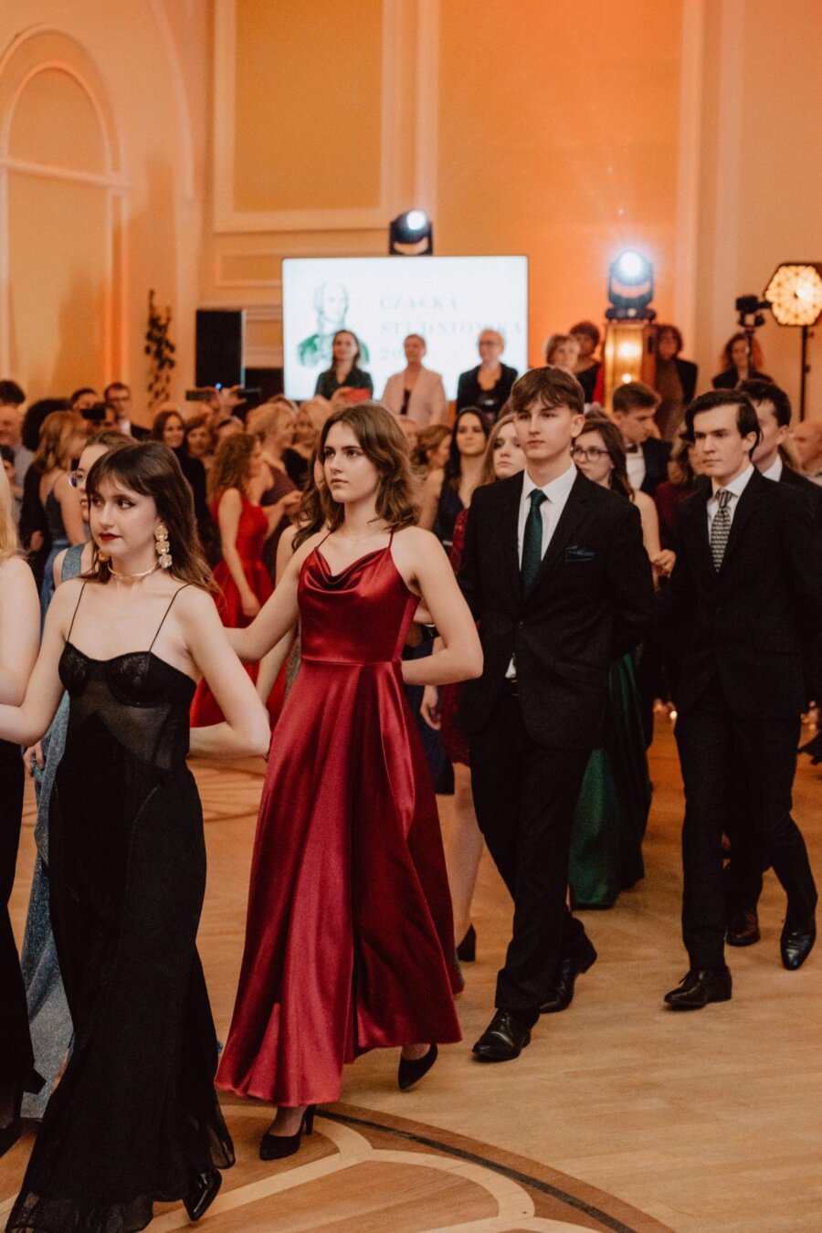 Grupa młodych ludzi ubranych formalnie, niektórzy w sukniach wieczorowych, inni w garniturach, spacerują parami po imprezie odbywającej się w pomieszczeniu. Sala jest elegancko urządzona, a w tle widoczny jest ekran. Atmosfera jest ożywiona i towarzyska – idealne miejsce na reportaż ze studniówki.