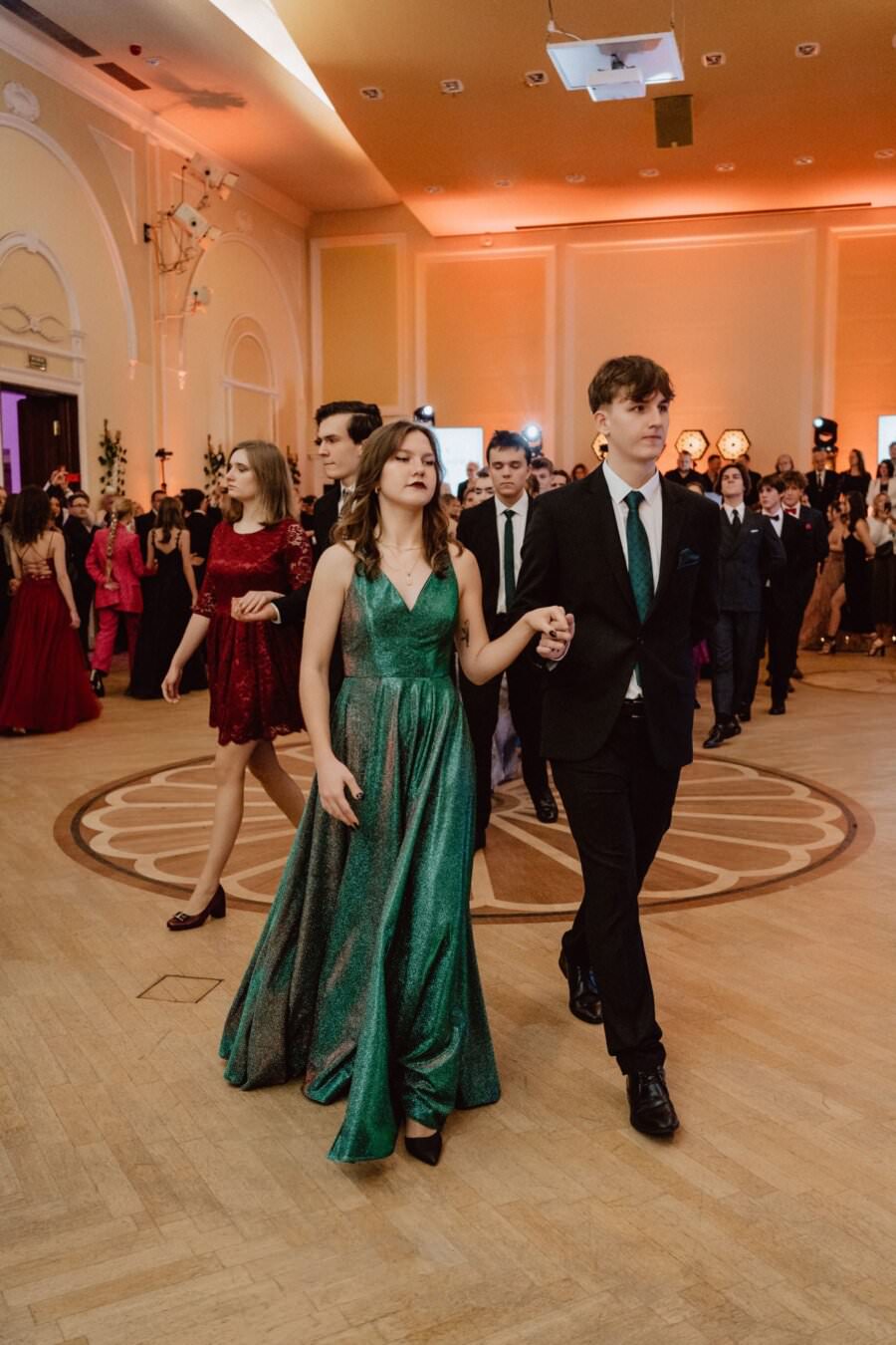 Grupa osób w sali balowej, elegancko ubranych, spacerujących w parach. Kobieta na pierwszym planie ma na sobie połyskującą zieloną suknię, a mężczyzna obok niej ma na sobie czarny garnitur z zielonym krawatem. W tle więcej par i ciepło oświetlone, bogate wnętrze, przypominające wybitny reportaż ze studniówki.