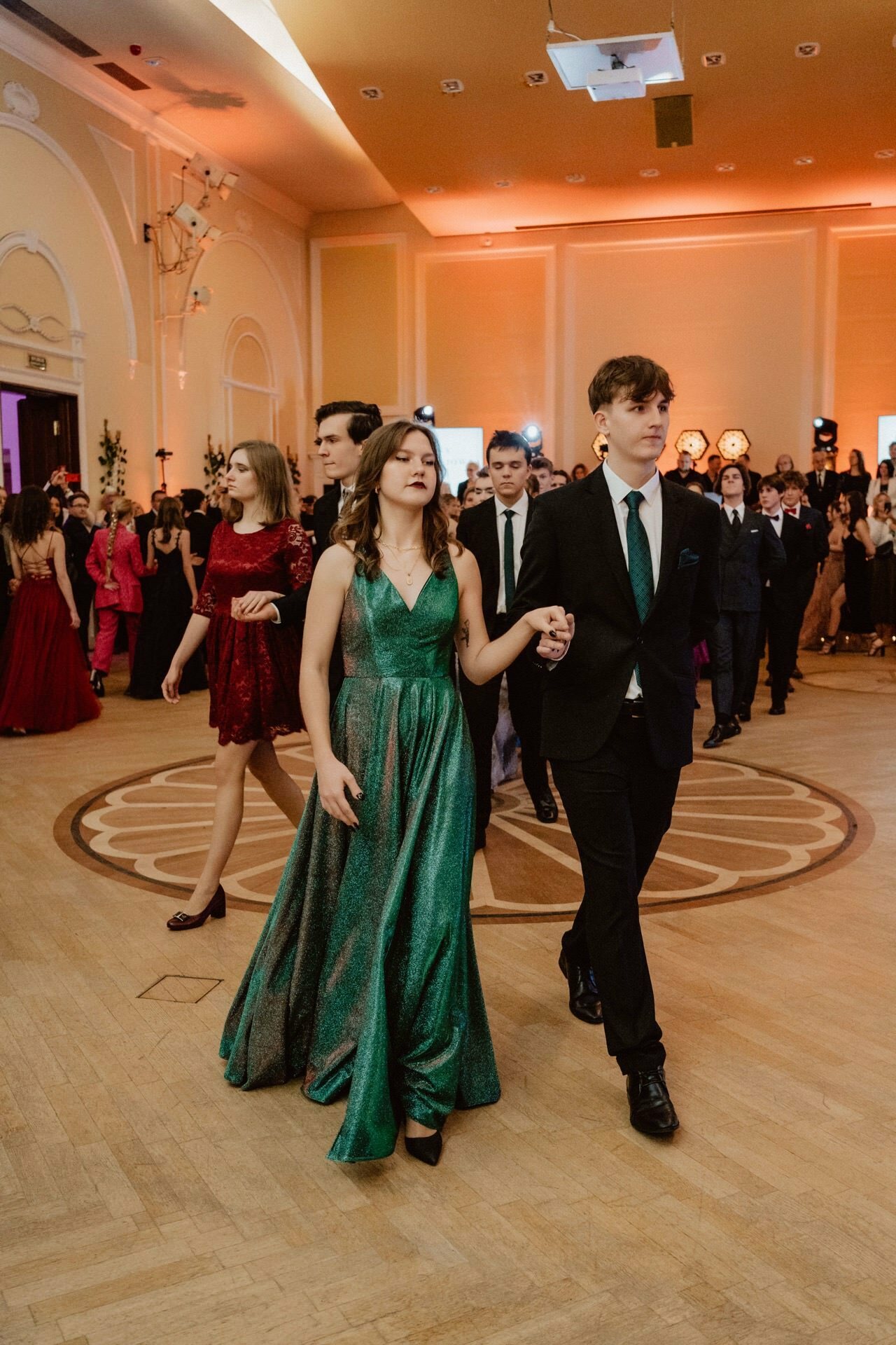 A group of people in a ballroom, elegantly dressed, walking in pairs. The woman in the foreground is wearing a shimmering green gown, while the man next to her is wearing a black suit with a green tie. In the background are more couples and a warmly lit, opulent interior, reminiscent of an outstanding prom reportage.  