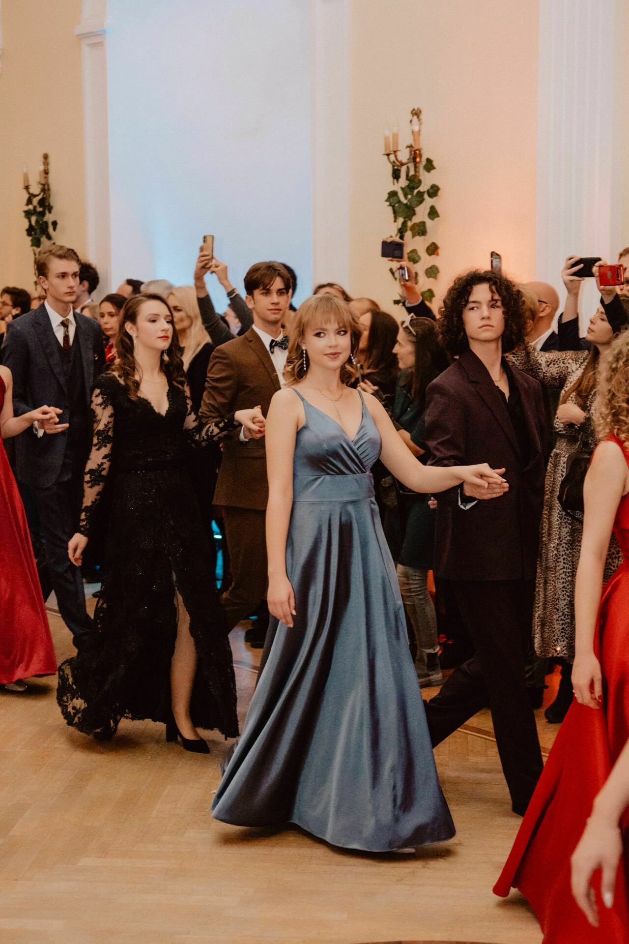 A group of elegantly dressed young adults walk in pairs at a formal event or ball, as if they were part of a prom reportage. They are surrounded by people watching and taking pictures. The woman in the foreground is wearing a blue dress, and the man accompanying her is in a dark suit. The atmosphere is festive.   