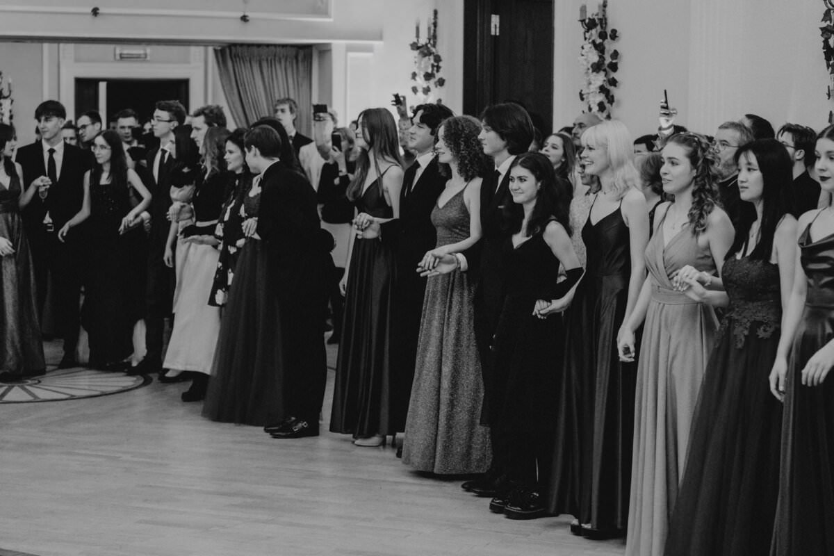 Czarno-białe zdjęcie grupy ludzi ubranych w formalne stroje, stojących w dużym pomieszczeniu o eleganckim wystroju. Niektórzy trzymają się za ręce lub łączą ramiona, podczas gdy inni robią zdjęcia telefonami. Wyglądają, jakby oglądali coś poza kamerą — ponadczasowy reportaż ze studniówki moment.
