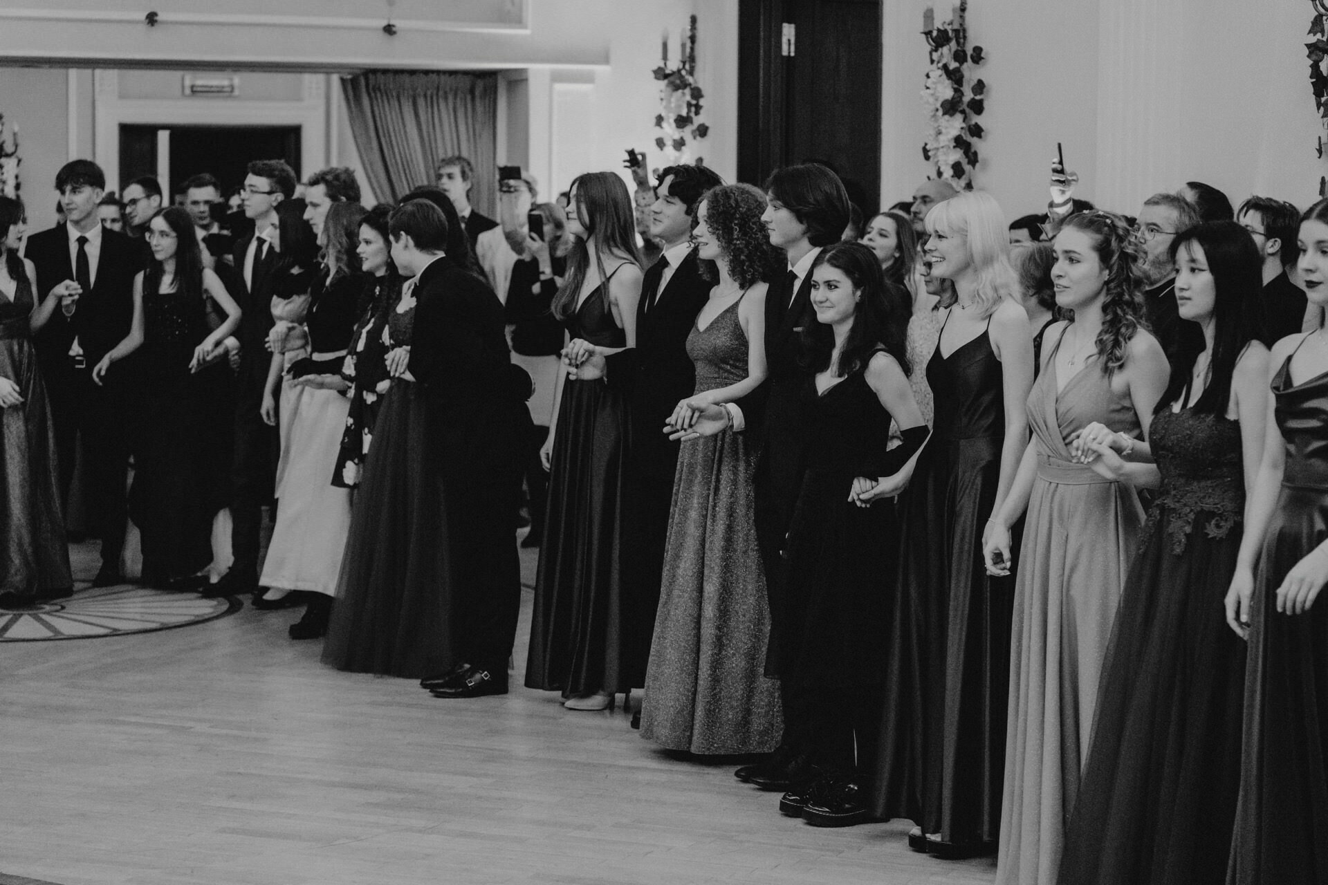 A black and white photo of a group of people dressed in formal attire, standing in a large room with elegant decor. Some are holding hands or linking arms, while others are taking pictures with their phones. They look as if they are watching something off-camera - a timeless reportage of a prom moment.  