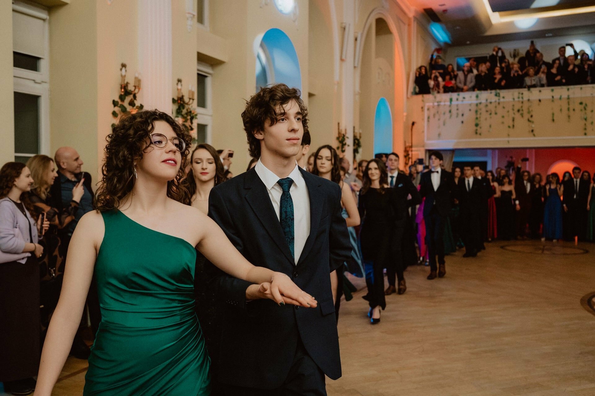 A young couple, elegantly dressed in formal attire, leads a procession at an event held indoors. The woman is dressed in a green one-shoulder dress, the man in a dark suit with a tie. The prom reportage captures the essence of the celebration, against the backdrop of other couples and onlookers.  