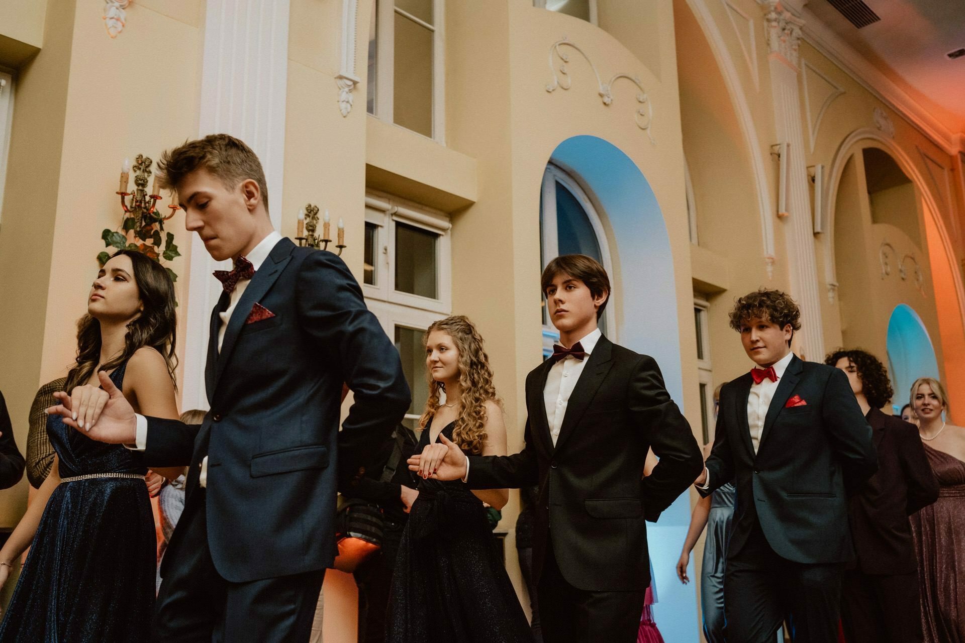 A group of young people dressed in formal attire walk in pairs. The men wear dark suits with red pillowcases, while the women wear elegant dresses. They look as if they are taking part in a report from a prom in the great hall.  