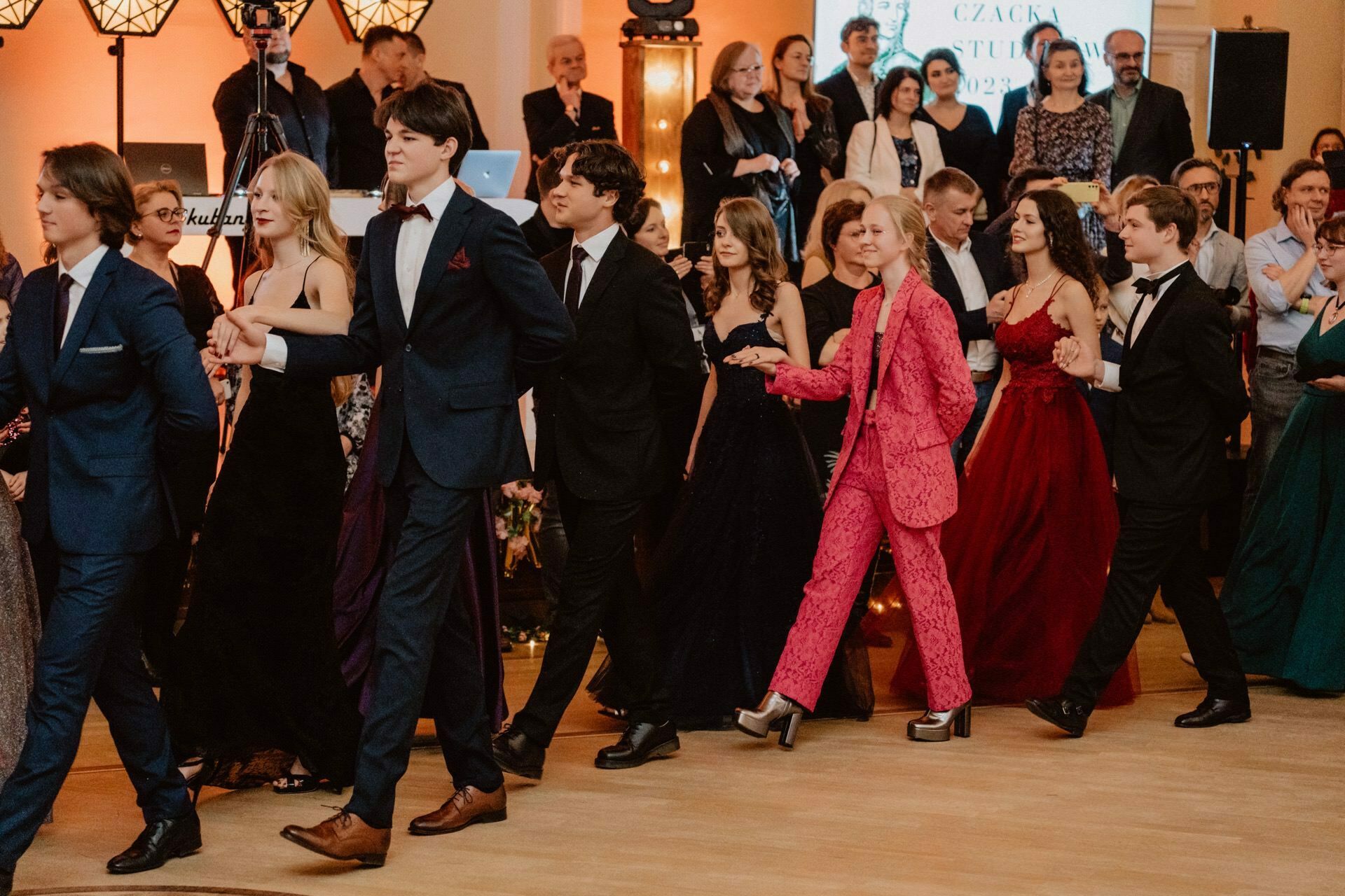 A group of young people dressed in formal attire dance in pairs during an elegant event reminiscent of a *prom prom*. The men wear suits and the women wear beautiful dresses. One person stands out in a bright pink suit. Spectators in the background are watching and taking pictures. The atmosphere is lively.    