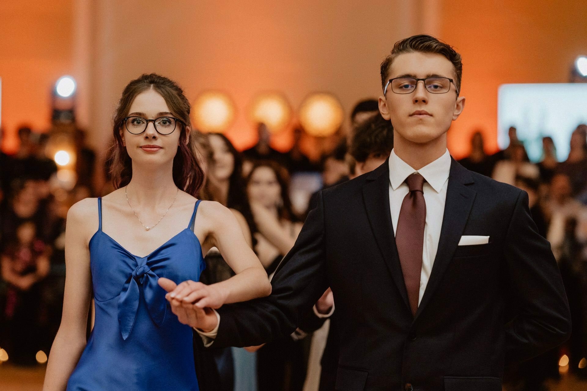 A young woman in a blue dress and a young man in a dark suit, white shirt and maroon tie dance together in a formal atmosphere. They hold hands and look ahead, while a crowd of people can be seen against a soft, orange-lit background, capturing the essence of a prom reportage. 