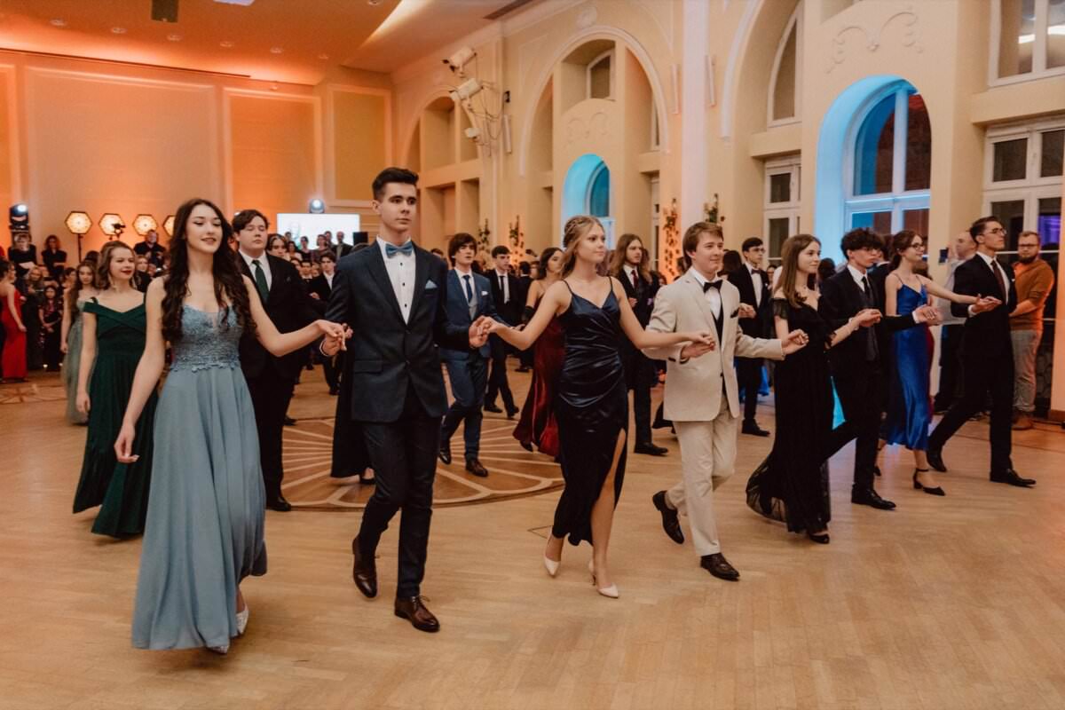 Grupa osób w strojach formalnych tańczy w parach w dużej, eleganckiej sali z łukowymi oknami i drewnianymi podłogami. Kobiety noszą długie suknie, a mężczyźni ubrani są w garnitury lub smokingi. Sala jest delikatnie oświetlona, co oddaje radosną atmosferę uroczystości „Reportażu ze studniówki”.