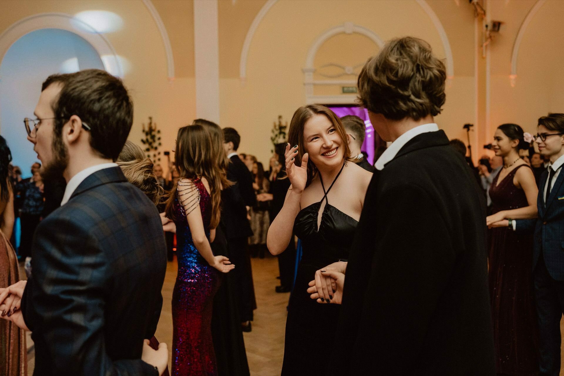 People in formal attire dance in a large, elegant hall with high, arched ceilings and ornate decor. A woman in a black dress smiles and holds hands with a man in formal attire as she dances. Other couples and groups also dance and talk in the background in this vibrant reportage of a prom.  