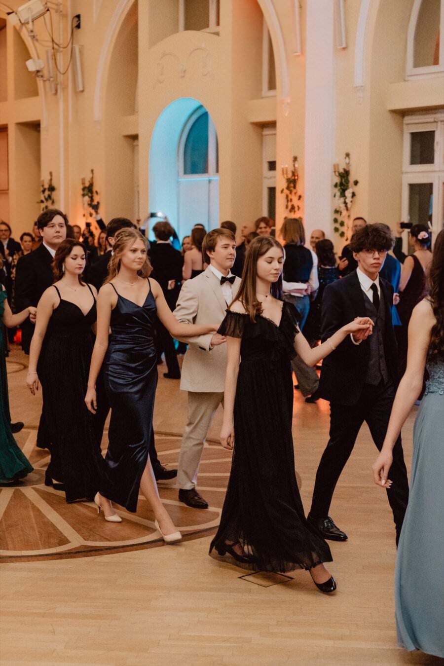 Grupa młodych ludzi ubranych formalnie w suknie i garnitury idzie parami po eleganckiej sali z łukowatymi drzwiami i wysokimi oknami. Wyglądają, jakby brali udział w formalnym wydarzeniu lub tańcu, przypominającym *reportaż ze studniówki*, ponieważ delikatnie oświetlona sala tworzy ciepłą atmosferę.
