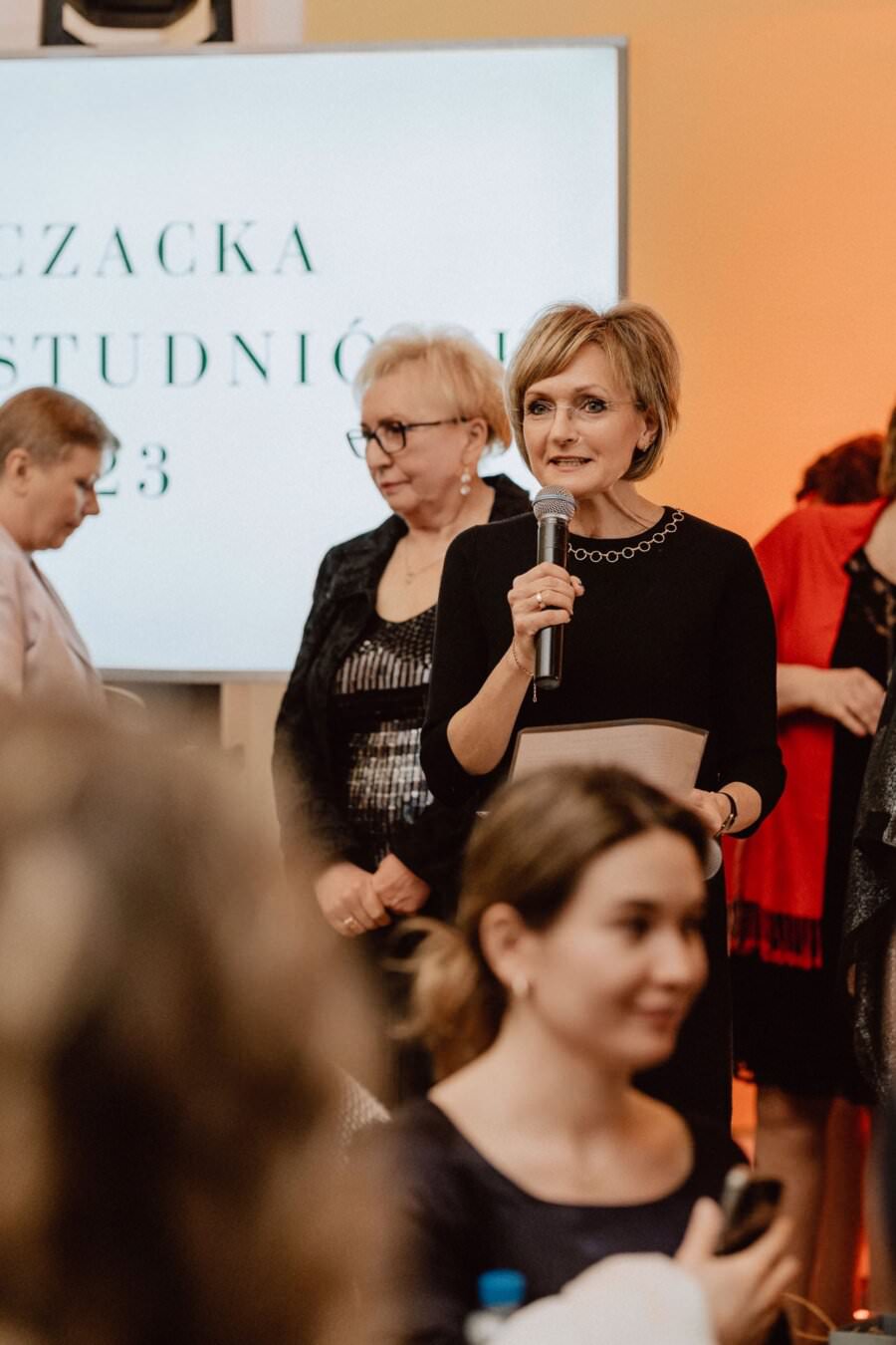 Kobieta w czarnej sukience przemawia do mikrofonu podczas imprezy odbywającej się w pomieszczeniach zamkniętych, relacjonując reportaż ze studniówki. Stoi na rozmytym tle z tekstem na ekranie. Wokół niej stoi kilka osób, głównie kobiet, jedna z nich siedzi i uważnie patrzy w stronę mówiącej.