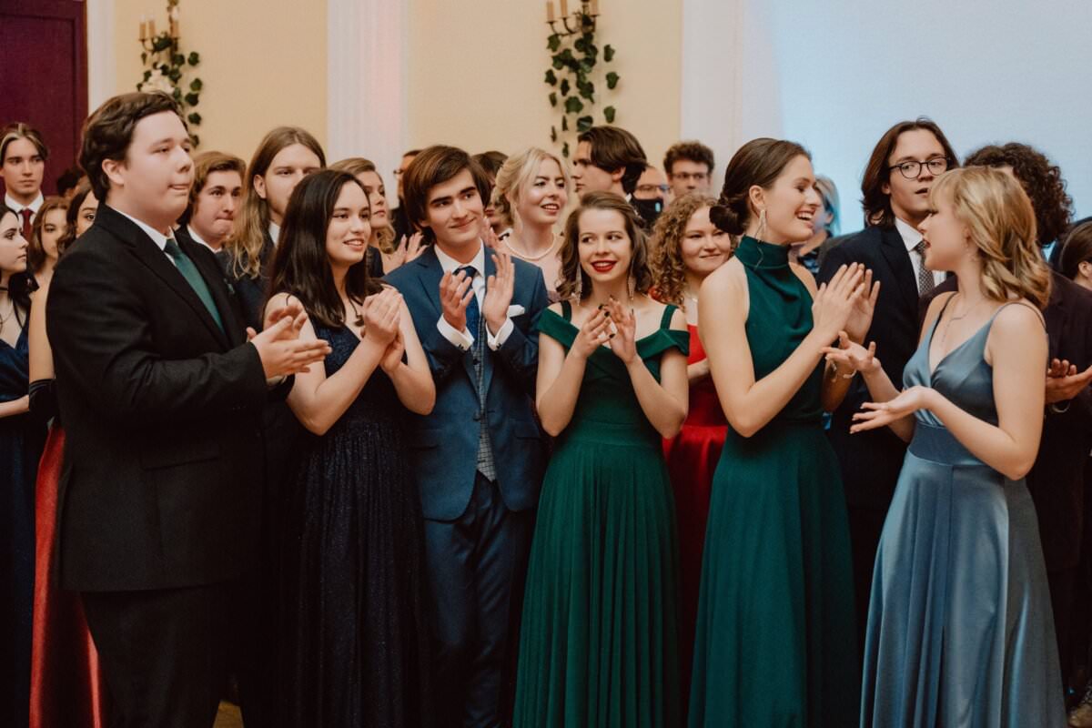 Grupa osób ubranych w stroje formalne stoi blisko siebie podczas imprezy odbywającej się w pomieszczeniu. Uśmiechają się i klaszczą, co sugeruje, że świętują lub cieszą się chwilą. Kilka osób ubranych jest w garnitury, inne w eleganckie suknie w różnych kolorach, co stanowi doskonały reportaż ze studniówki.