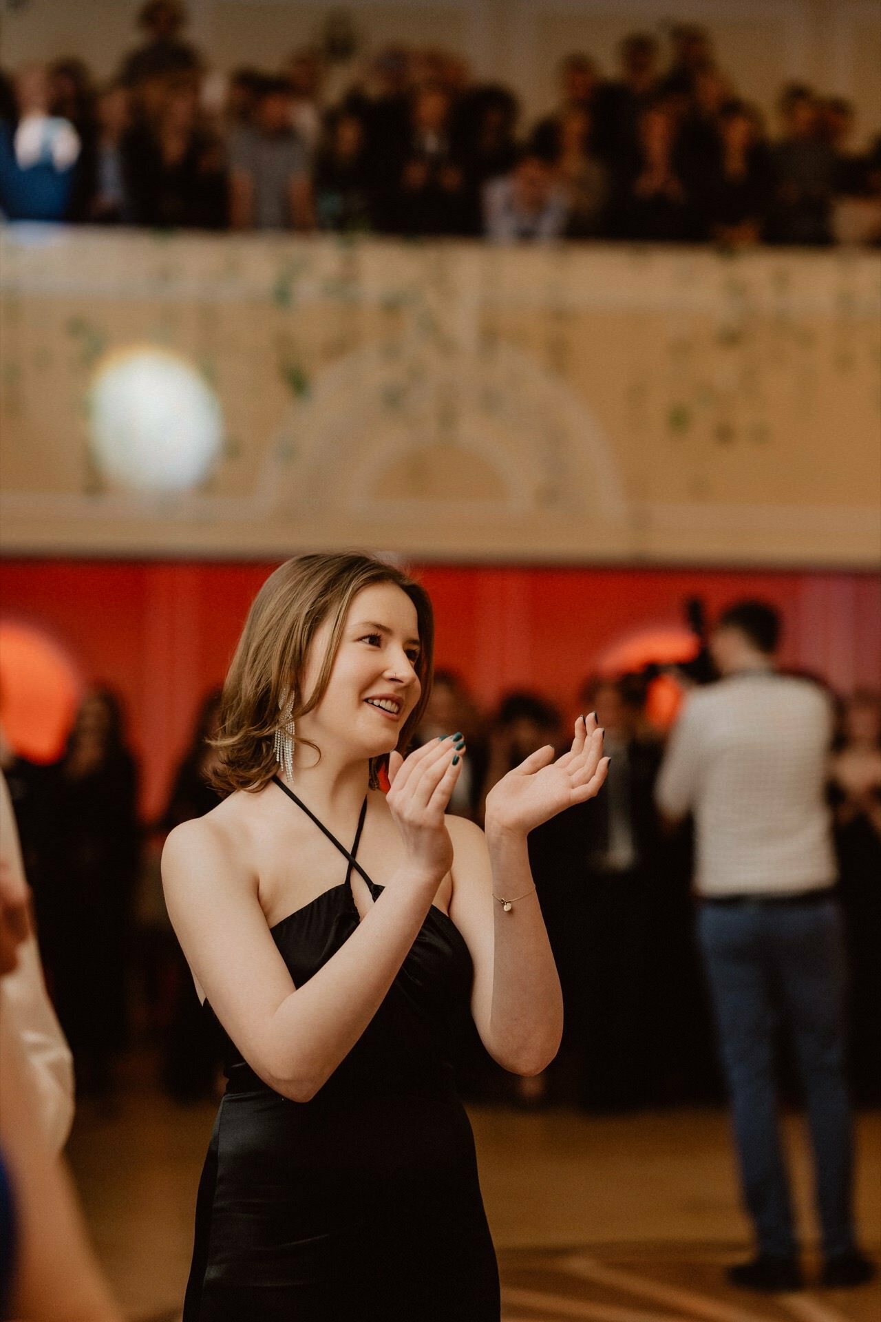 A woman in a black dress claps and smiles as she stands in a large hall, reminiscent of a prom reportage. In the background you can see a blur of people, some on a higher floor and others on the same floor as the woman. The place looks like an event or celebration.  