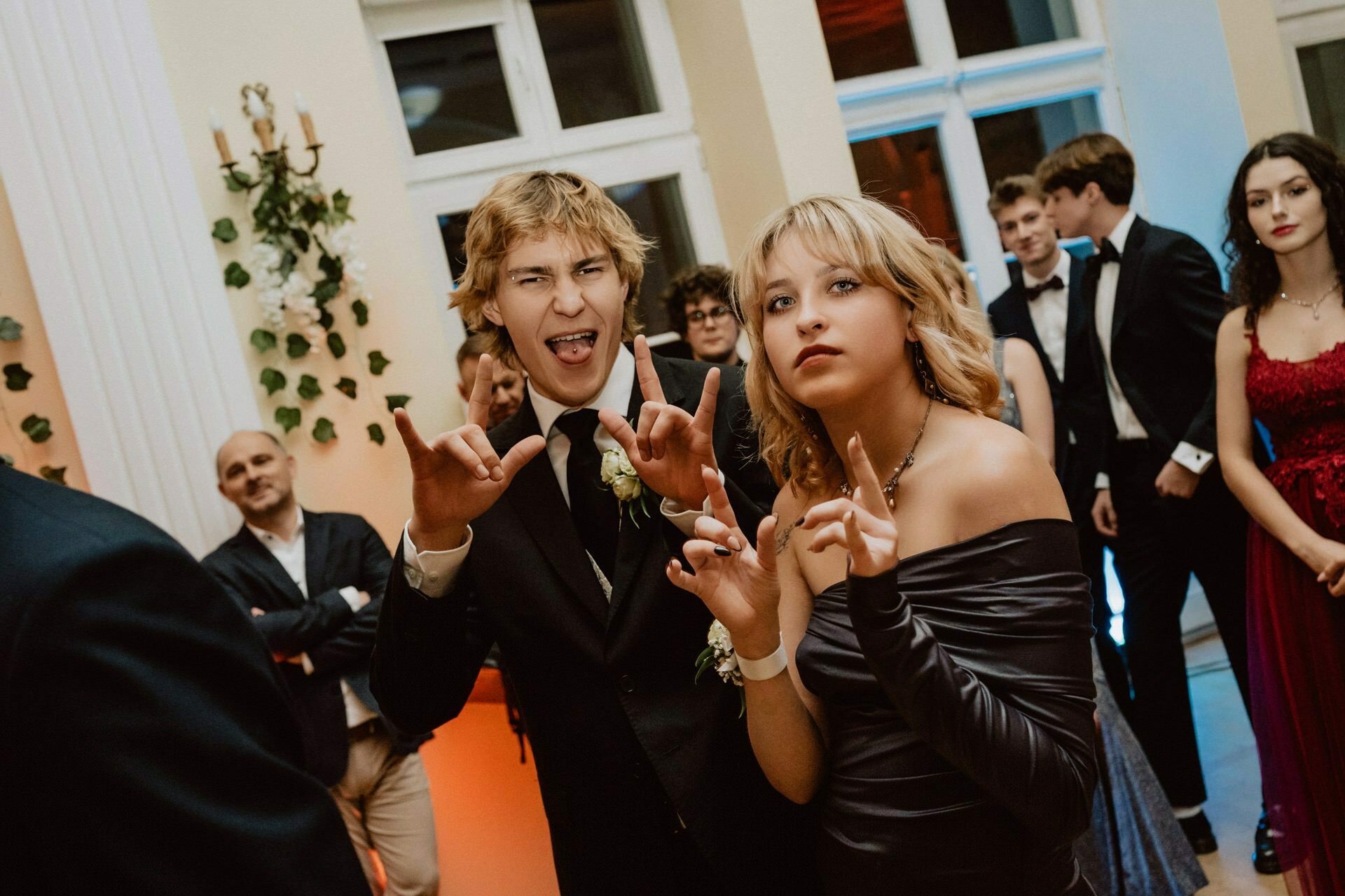 Two young adults at an official event pose playfully for the camera. The young man makes a rock and roll gesture with both hands, while the young woman next to him also takes a playful pose. Both are dressed in formal attire, probably part of a reportage from a prom, and other participants can be seen in the background.  