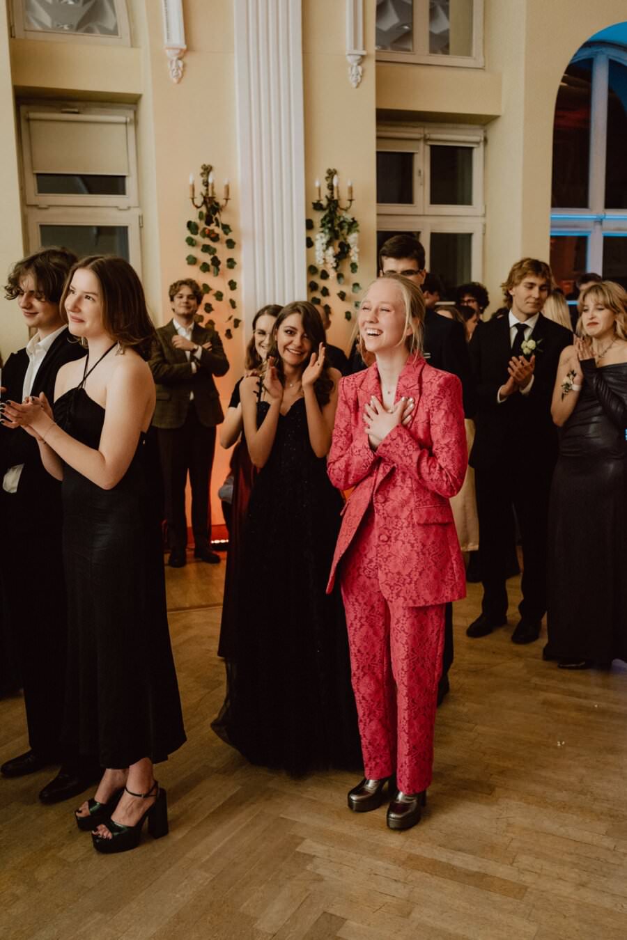 Grupa osób ubranych formalnie stoi w dobrze oświetlonym pomieszczeniu ozdobionym zielenią. Niektórzy klaszczą, uśmiechają się i patrzą w stronę niewidzialnego wydarzenia. Jedna osoba na pierwszym planie wyróżnia się w jasnoróżowym koronkowym kostiumie, wyrażając podekscytowanie i radość — tętniący życiem moment uchwycony niczym *reportaż ze studniówki*.