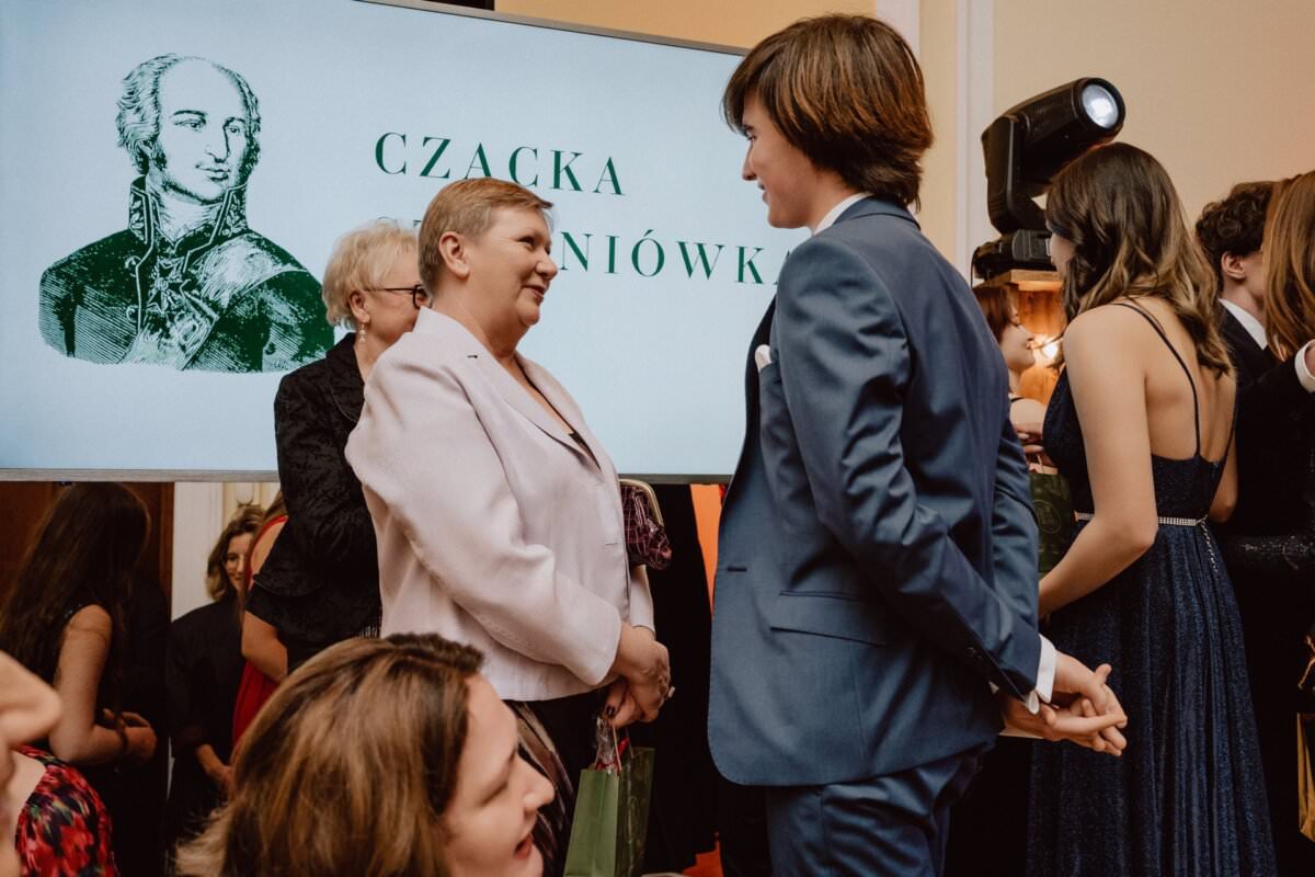 Grupa ludzi na oficjalnym wydarzeniu, niektórzy w garniturach i sukienkach. Rozmową zajmują się dwie osoby, jedna w garniturze, a druga w jasnym stroju. W tle ekran wyświetlający tekst i ilustrację postaci historycznej, przypominający reportaż ze studniówki.