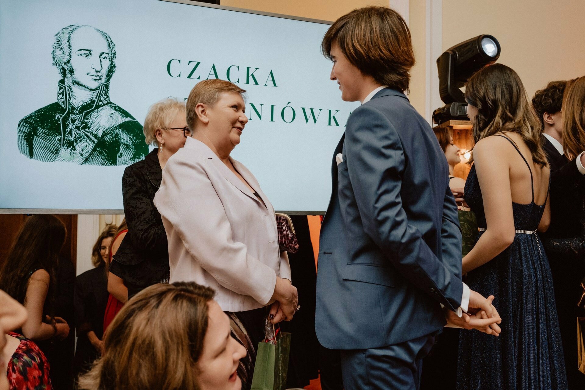 A group of people at a formal event, some in suits and dresses. Two people, one in a suit and the other in a bright outfit, are engaged in conversation. In the background is a screen displaying text and an illustration of a historical figure, reminiscent of a report from a prom.  