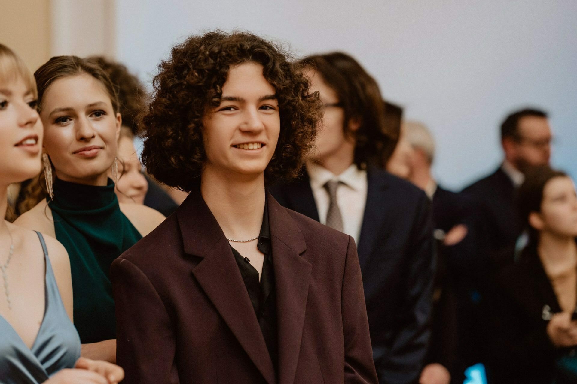 A young person with curly hair, wearing a dark jacket, smiles at the camera while standing among a group of people in what looks like a reportage from a prom. Two people in elegant attire are visible in the background, one in a green dress, the other in a suit and glasses. 