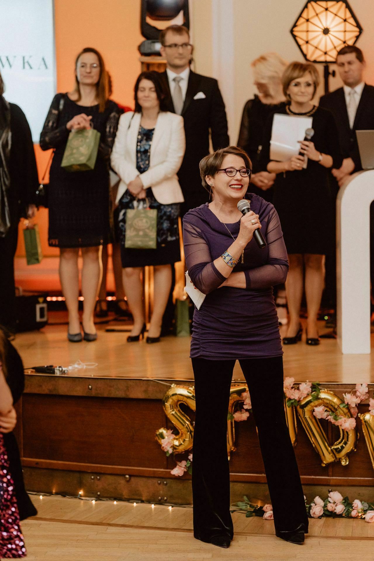 In the foreground of the picture stands a person, holding a microphone and smiling, speaking to the group. Dressed in a purple top and dark pants, they stand on a decorated stage. Behind them stand several formally dressed people, holding gift bags - a vibrant scene straight out of a prom report.  