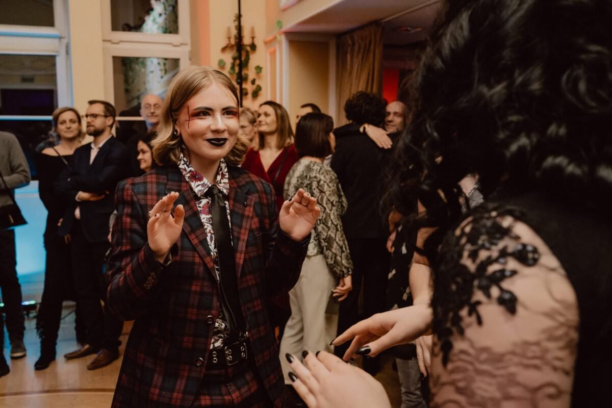 Osoba w czerwono-czarnym garniturze w kratę uśmiecha się i podnosi ręce podczas tańca na imprezie towarzyskiej. W tle mieszają się inni uczestnicy, ubrani w stroje wizytowe. Reportaż ze studniówki oddaje ożywioną, świąteczną atmosferę, a dekoracje dodają jej uroku.