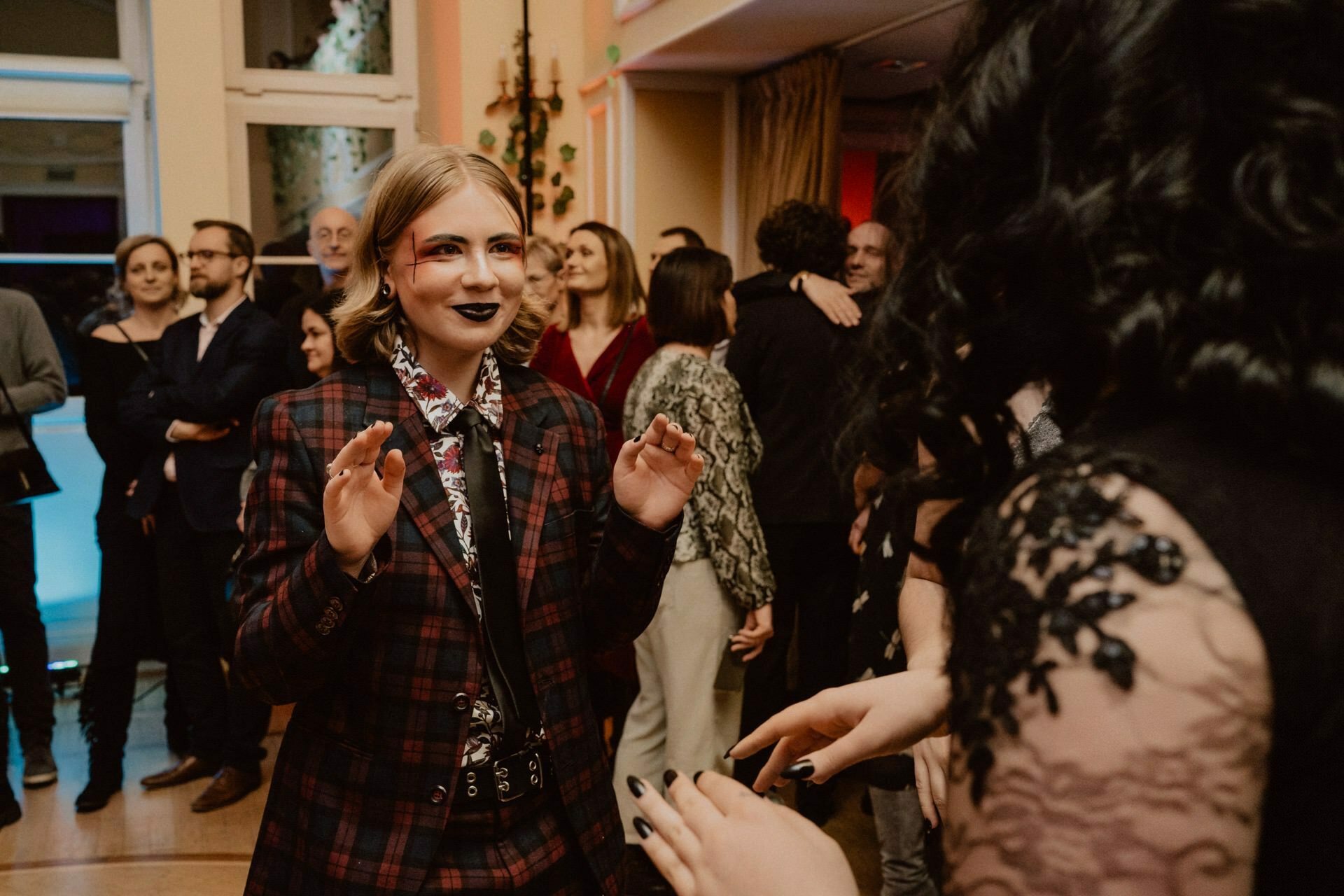 A person in a red and black plaid suit smiles and raises his hands while dancing at a social event. Other attendees, dressed in formal attire, mingle in the background. The reportage of the prom captures the lively, festive atmosphere, and the decorations add to its charm.  