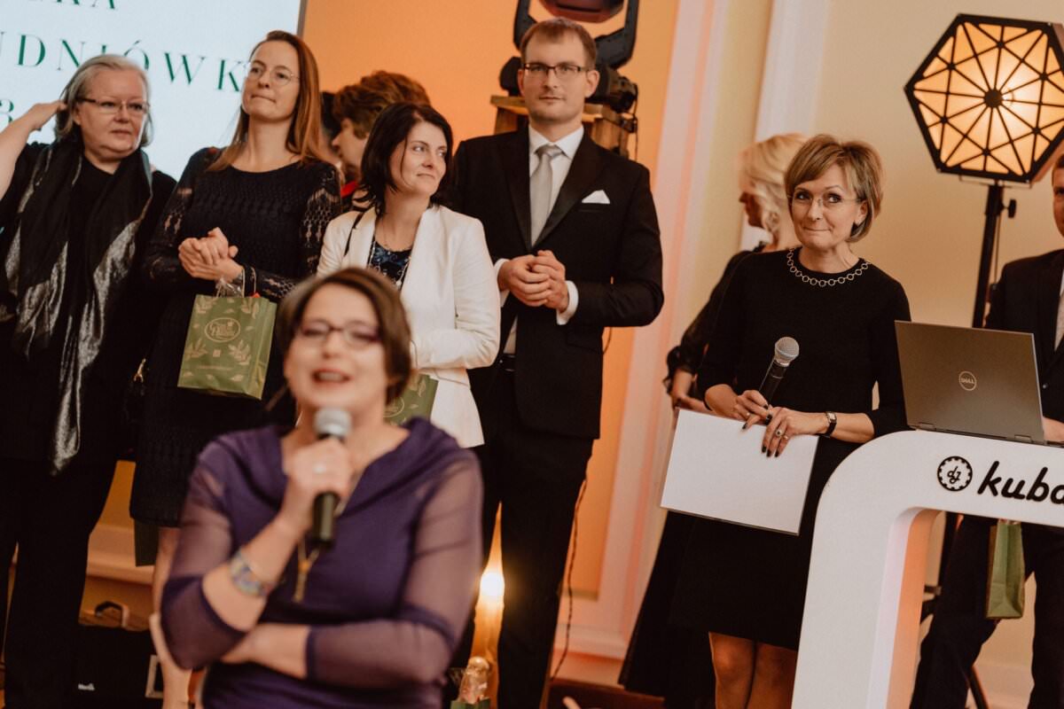 Grupa osób ubranych w stroje formalne stoi razem w dobrze oświetlonym pomieszczeniu. Niektórzy trzymają papiery, jedna kobieta mówi do mikrofonu. Na pierwszym planie widoczny laptop na podium. W tle duży ekran wyświetla częściowo widoczny tekst – oddając istotę „reportażu ze studniówki.