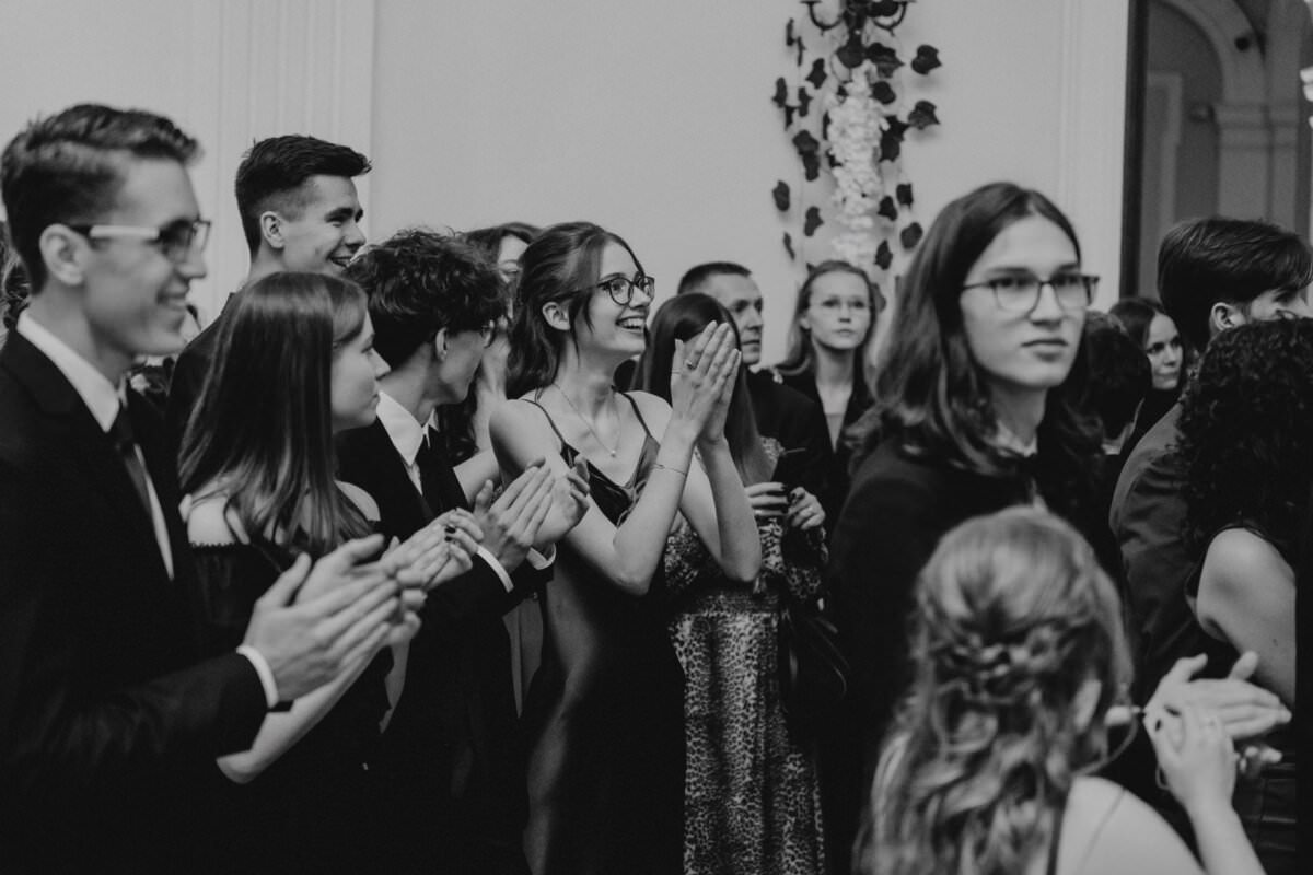 Czarno-białe zdjęcie przedstawiające grupę ludzi ubranych formalnie, stojących blisko siebie w pokoju. Wygląda na to, że klaszczą i wiwatują, niektórzy uśmiechają się i wyglądają na podekscytowanych. Kobieta w centrum klaszcze w dłonie, emanując radością – oddając esencję reportażu ze studniówki.