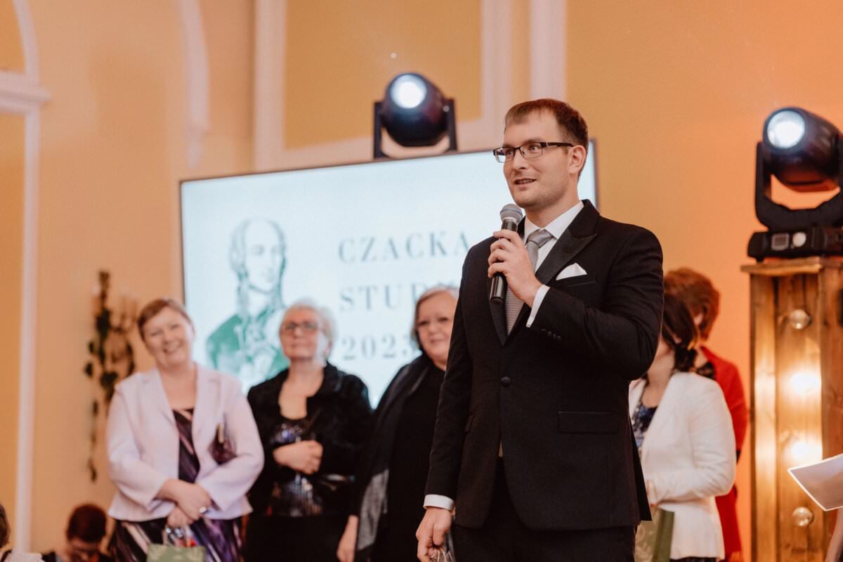 Podczas wydarzenia mężczyzna w czarnym garniturze i krawacie przemawia do mikrofonu. Za nim pięć osób stoi i uśmiecha się. Na ekranie w tle widoczny jest portret oraz częściowo widoczny napis „CZACKA STU…” wskazujący, że jest to reportaż ze studniówki. Widoczne są także światła sceniczne.