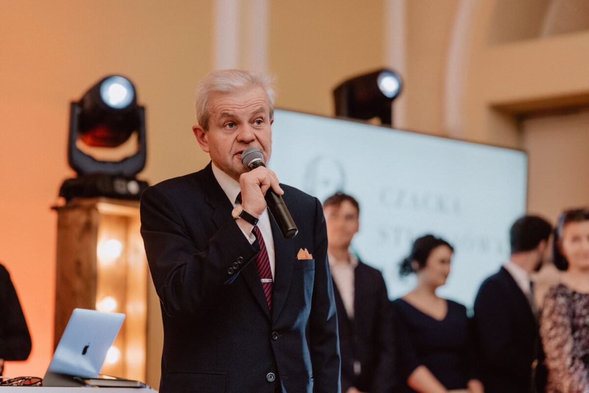 Do mikrofonu przemawia starszy mężczyzna o siwych włosach, ubrany w ciemny garnitur i krawat w paski. Za nim rozmyte tło z kilkoma osobami i ekranem, przypominające klimat „reportażu ze studniówki”. Miejsce wydaje się być formalnym wydarzeniem lub prezentacją.