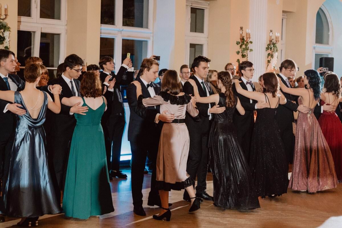 Grupa elegancko ubranych osób tańczy w parach w formalnej sali balowej, przypominającej reportaż ze studniówki. Mężczyźni w smokingach i kobiety w wieczorowych sukniach wirują i wykonują skoordynowane ruchy taneczne. Sala jest ozdobiona dużymi oknami i gustownymi dekoracjami.
