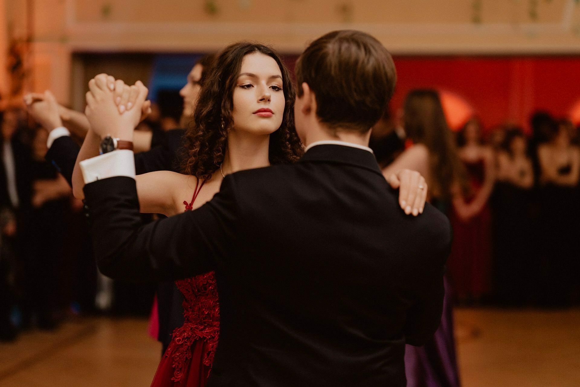 A couple dressed in formal attire dance closely at a ballroom party. The woman with curly hair is wearing a red dress and looks poised as she holds the man's hand. The man, with his back turned to the camera, is dressed in a black suit. Other dancing couples can be seen in the background, capturing the essence of the prom reportage.   