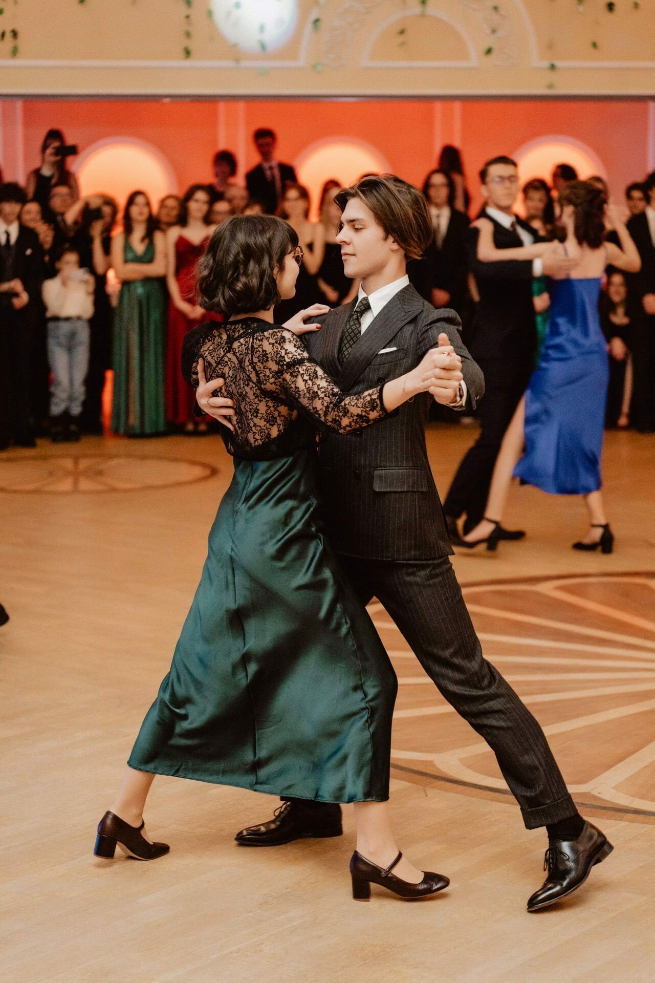 A young couple dances elegantly together at a formal event. The woman is dressed in a green dress and black stilettos, the man is dressed in a dark suit and black shoes. In the background, spectators in formal attire watch them, giving the impression of a scene from a prom reportage.  