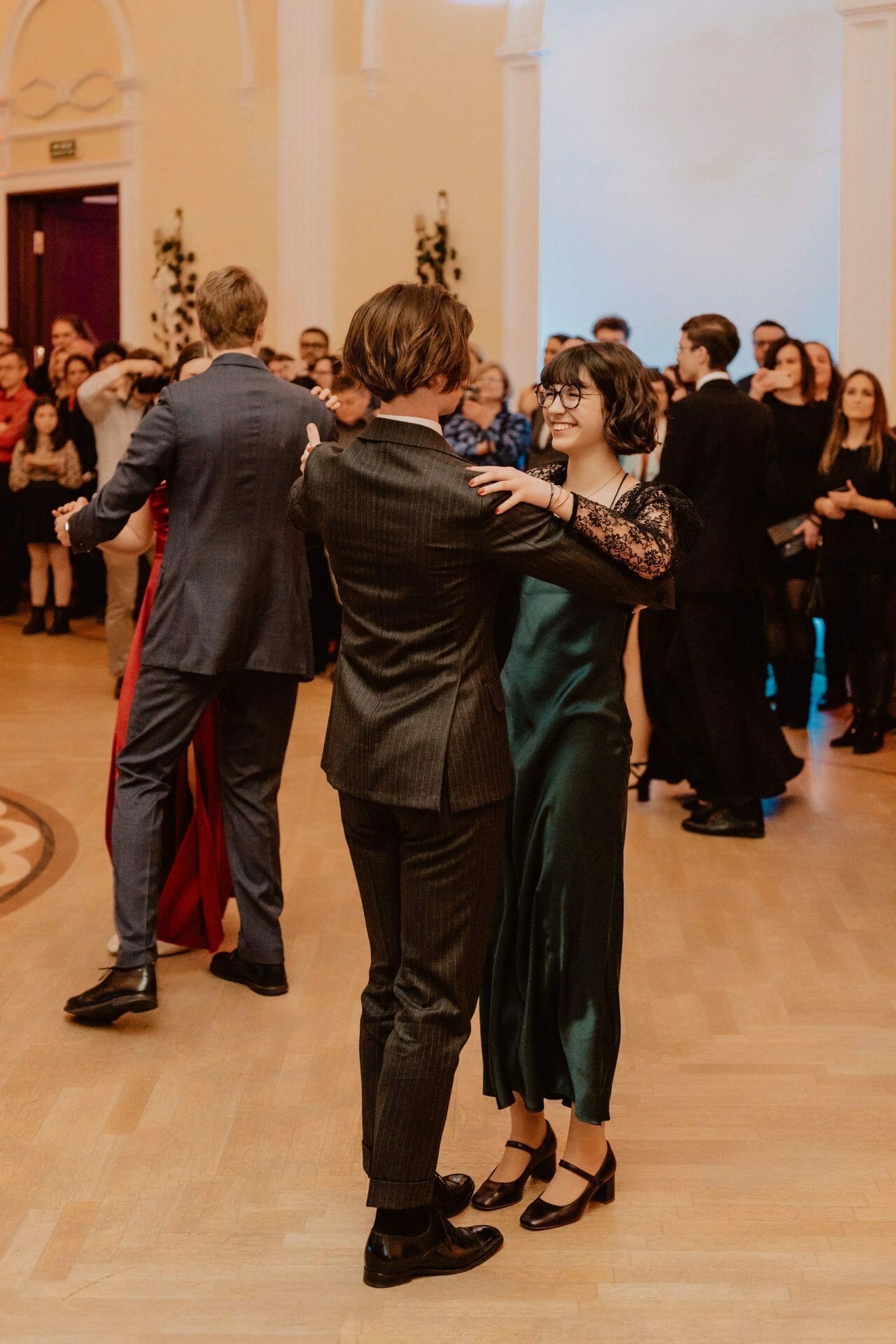 A couple dances gracefully in the foreground during an elegant event, reminiscent of a prom reportage, with a group of people in the background dancing or watching. The room features classic architecture with tall columns and white walls, creating a sophisticated atmosphere. 