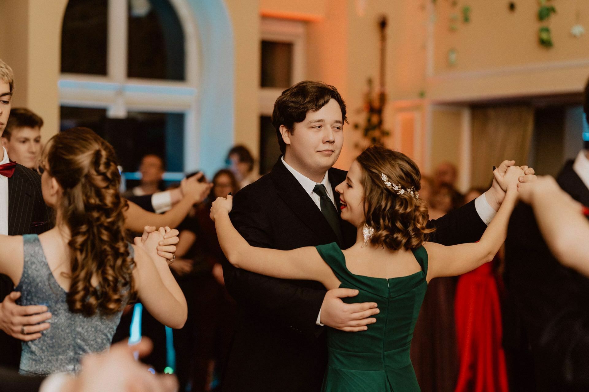 A young man and woman, both dressed in formal attire, are dancing together in a well-lit room filled with other couples. The woman is wearing an emerald green dress and a floral hair ornament, the man is wearing a black suit. The other participants are visible in the background, capturing the essence of the prom reportage.  