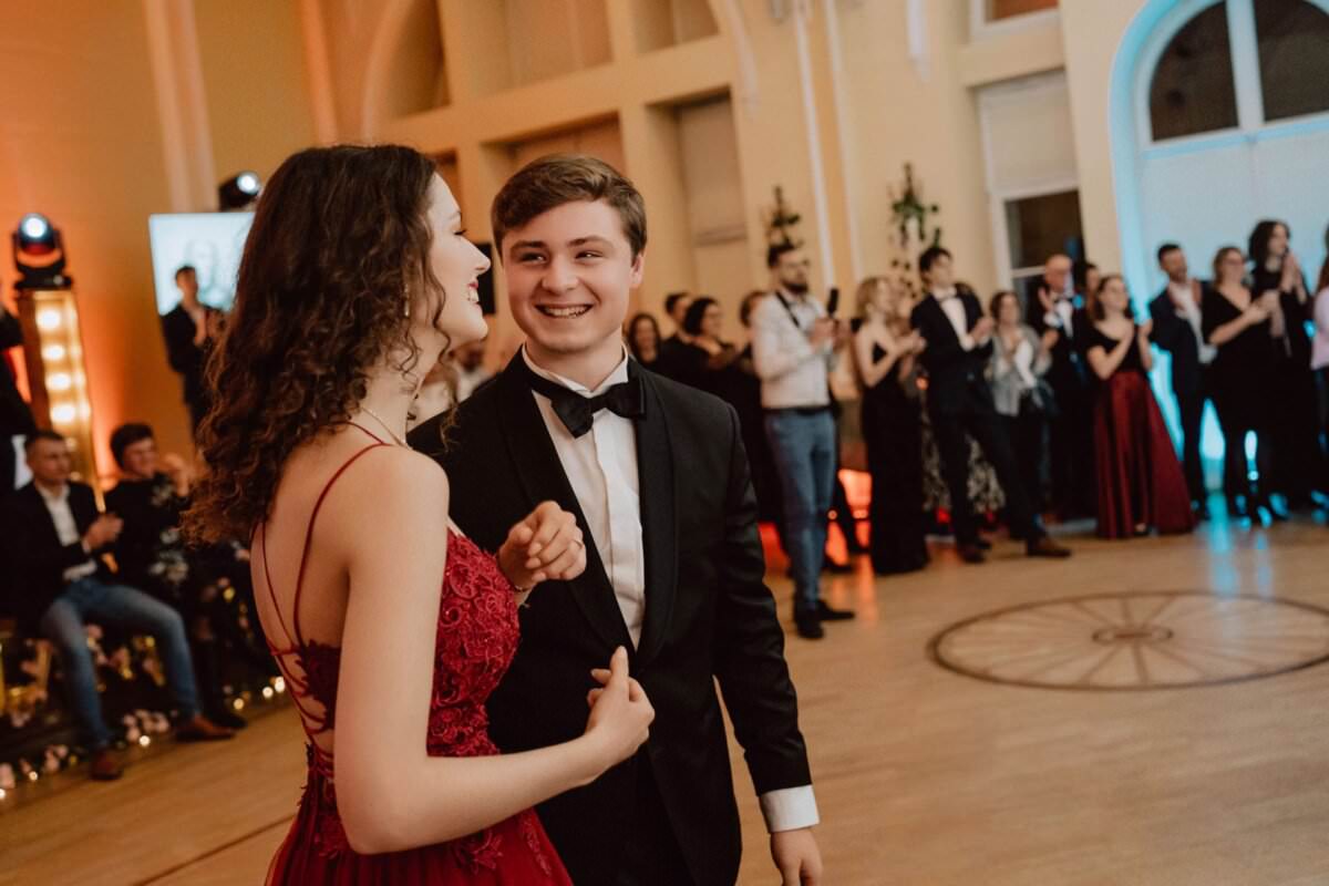 Młoda para, ubrana formalnie, tańczy i dzieli radosne chwile na fantazyjnej imprezie odbywającej się w pomieszczeniu. Kobieta ma na sobie czerwoną sukienkę, a mężczyzna czarny garnitur z muszką. W tle obserwują i klaszczą pozostali goście, także w strojach formalnych – idealna scena na reportaż ze studniówki.