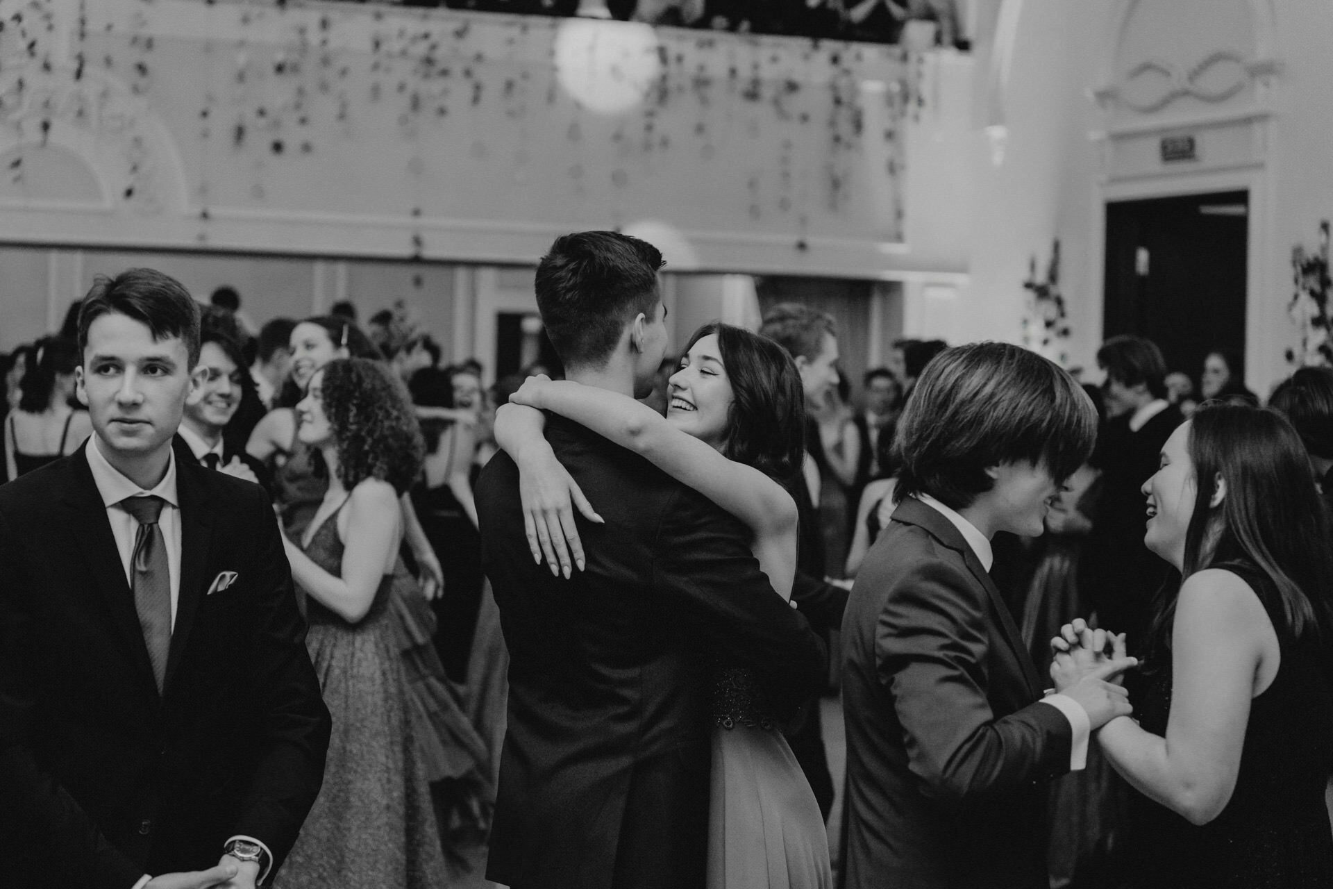 Black and white photo showing a formal dance, reminiscent of a reportage from a prom. The couple in the middle embrace each other surrounded by other couples dancing. People in formal attire, some smiling and talking. The venue has elegant decor, with a visible balcony and wall decorations.   