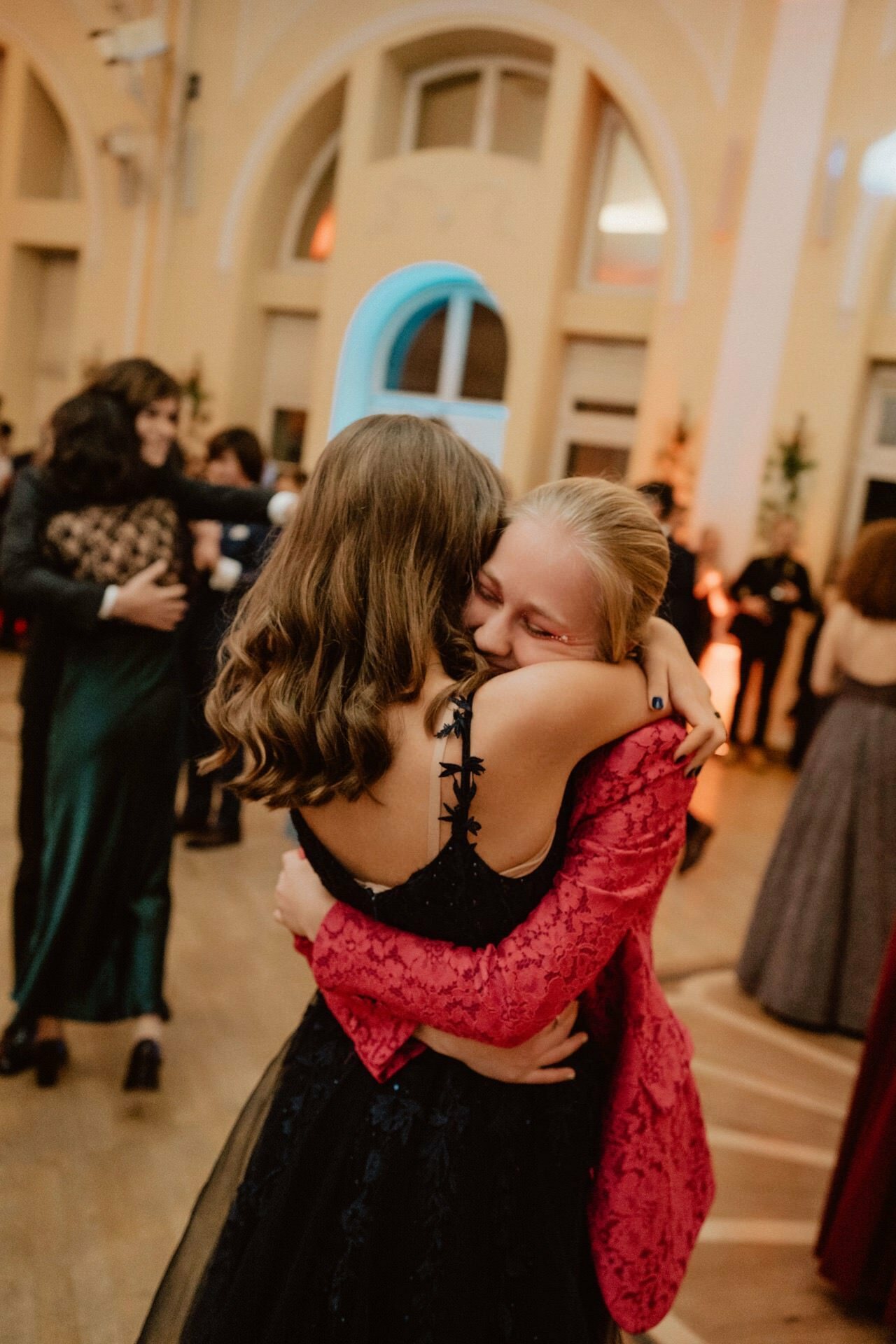 Two people embrace each other tightly at an official event and smile warmly at each other. The person on the left has long brown hair and is wearing a black dress, while the person on the right has blonde hair tied back and wears a red lace dress. In this reportage from the prom, other guests mingle in the background.  