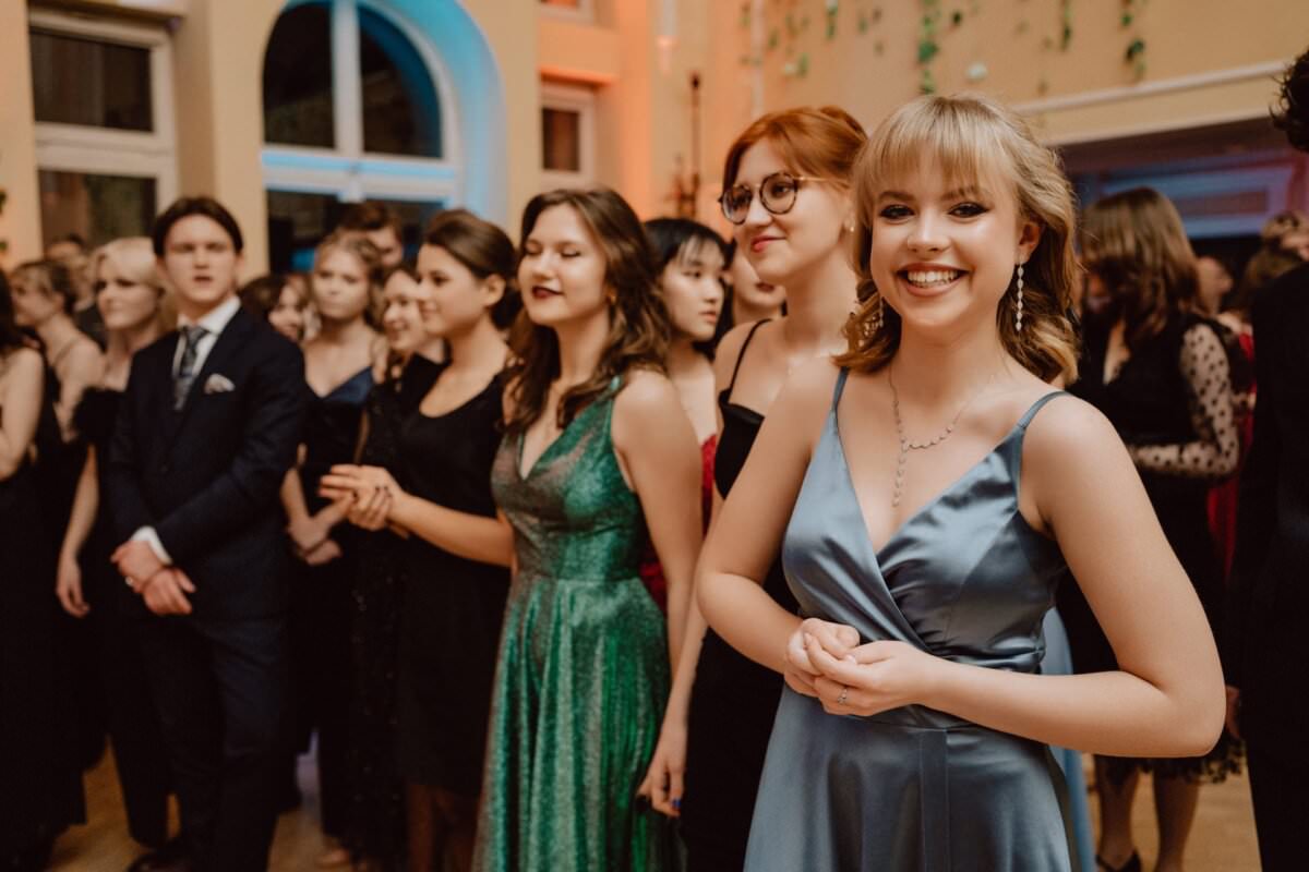 Grupa osób w strojach formalnych stoi w udekorowanym pokoju. Na pierwszym planie uśmiecha się młoda kobieta w niebieskiej satynowej sukience, a inne za nią, ubrane w różne sukienki i garnitury, prowadzą rozmowę. Pomieszczenie ma łukowate okna, a na ścianach wiszą pasma zieleni – reportaż ze studniówki doskonale rejestruje każdą chwilę.