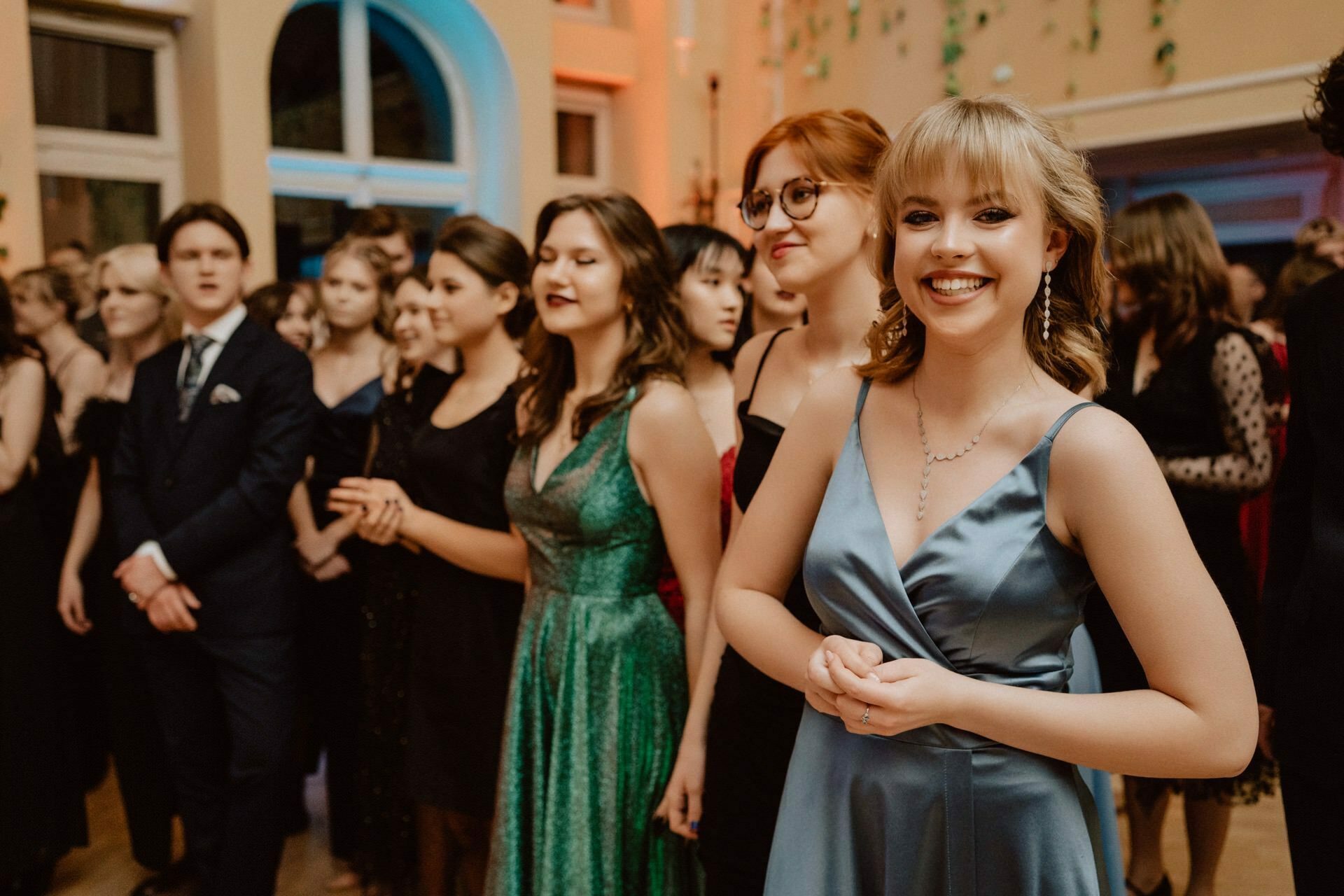 A group of people in formal attire stand in a decorated room. A young woman in a blue satin dress smiles in the foreground, while others behind her, dressed in various dresses and suits, carry on a conversation. The room has arched windows, and strands of green hang on the walls - the reportage of the prom perfectly captures every moment.  