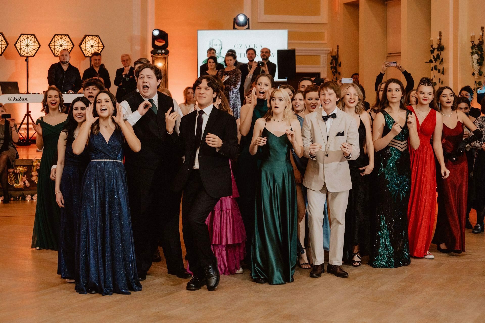 A group of young adults dressed in formal evening wear are standing in a ballroom. They look excited and cheer or applaud. The backdrop is a musical band, stage and decorative lighting, capturing the festive mood of the prom reportage.  