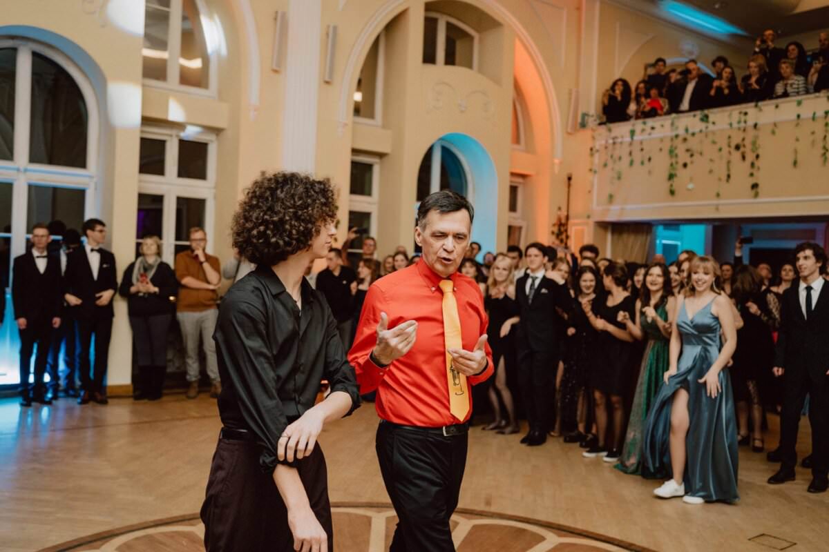 Dwie osoby stoją na środku dużego, elegancko urządzonego pokoju i prowadzą rozmowę. Jeden ma na sobie czarną koszulę, drugi jaskrawoczerwoną koszulę z żółtym krawatem. Otacza ich tłum uśmiechniętych, formalnie ubranych ludzi, niektórzy tańczą, inni patrzą – prawdziwy reportaż ze studniówki.
