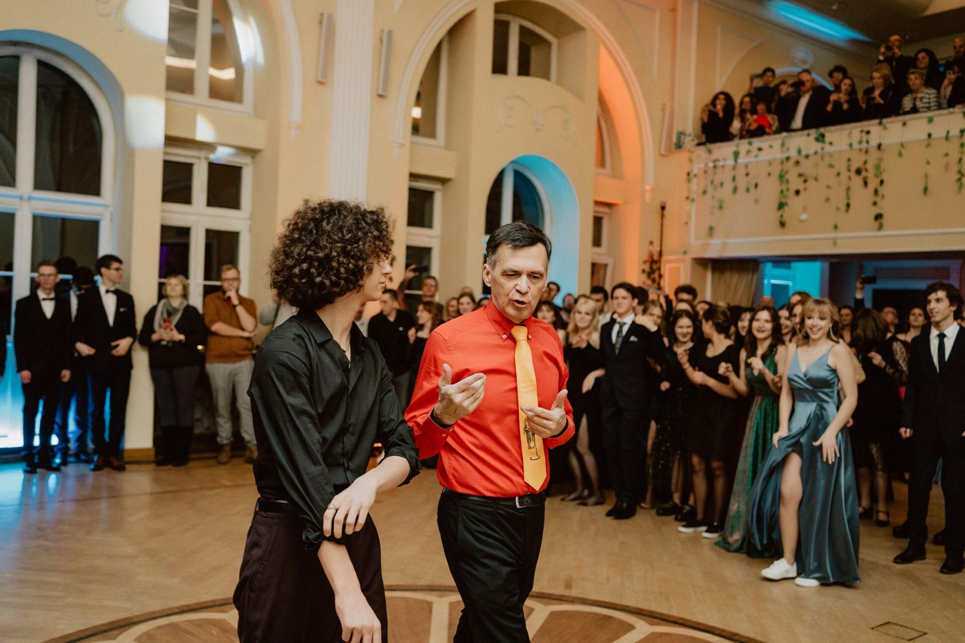 Two people are standing in the middle of a large, elegantly decorated room having a conversation. One is wearing a black shirt, the other a bright red shirt with a yellow tie. They are surrounded by a crowd of smiling, formally dressed people, some dancing, others looking on - a real reportage from a prom.  